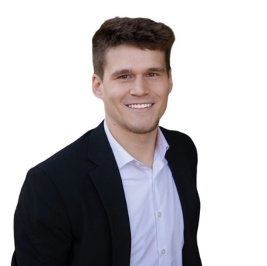 Portrait of a young man with brown hair wearing a dark blazer and white shirt, smiling against a white background.