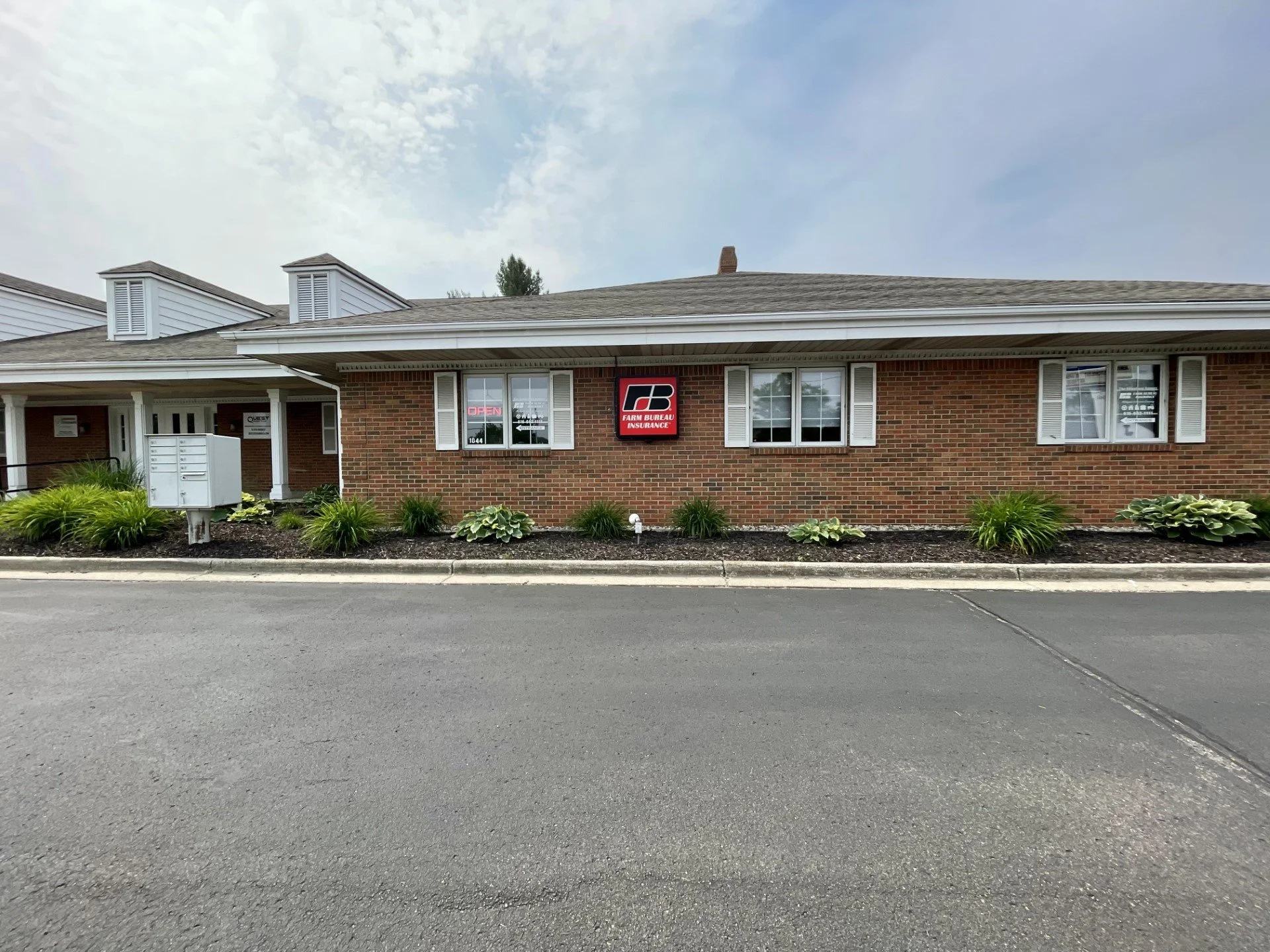 A brick commercial building with a sign that reads 'Farm Bureau Insurance' and a neon 'Open' sign in the window. There is a landscaped area with green plants and a row of mailboxes in front.
