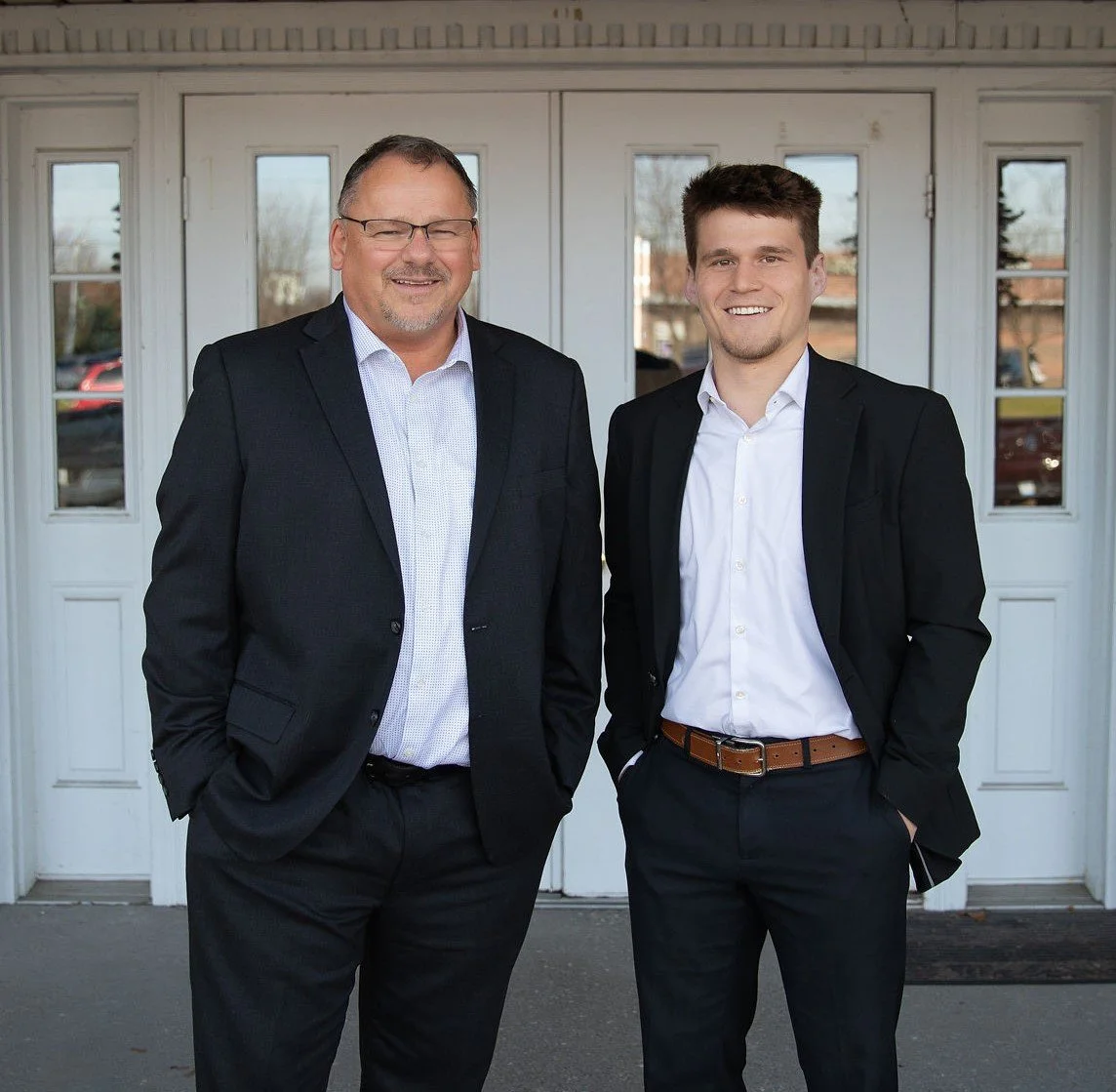 Two men in suits standing outside in front of a white building with glass-paneled doors, smiling at the camera.