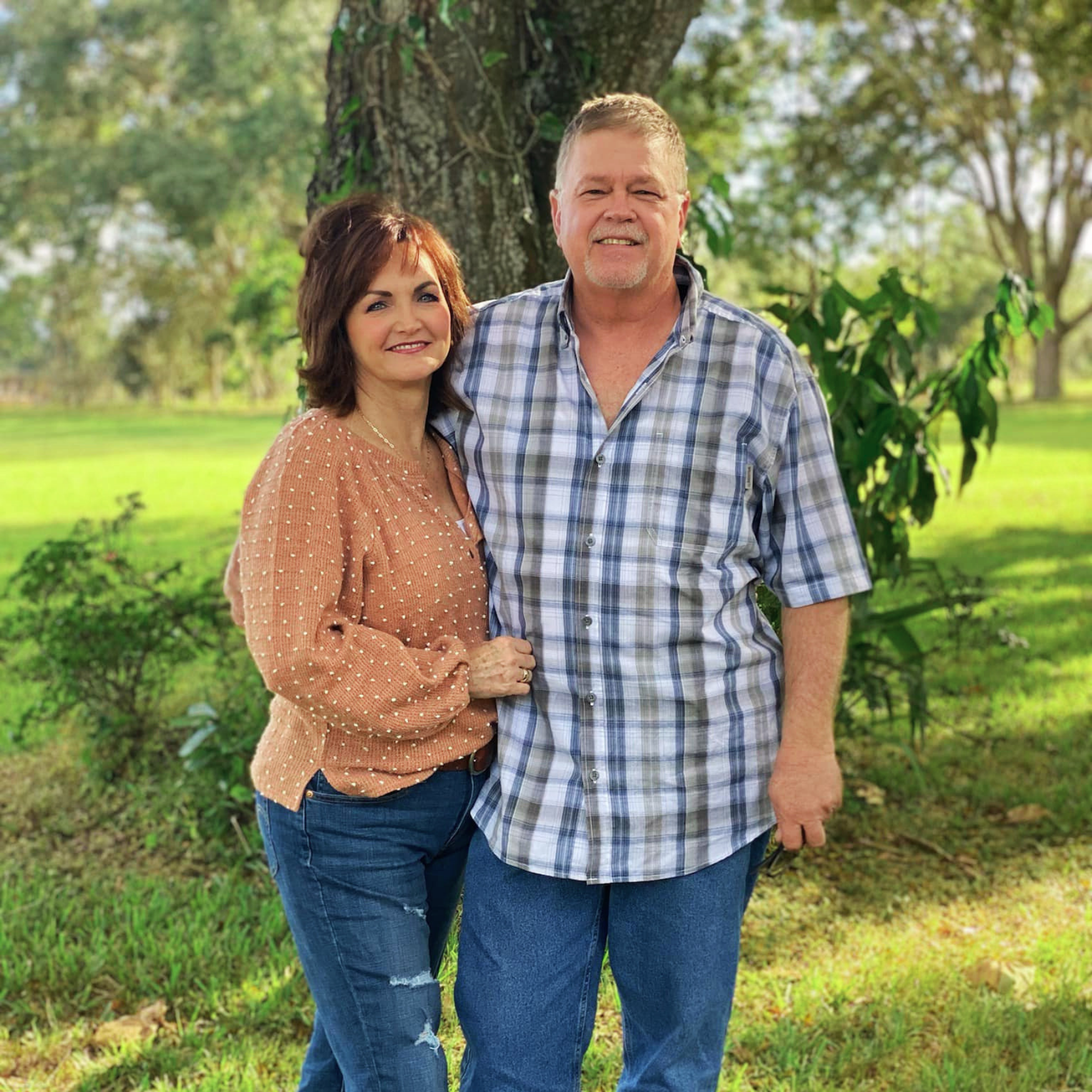 A smiling couple standing outdoors by a tree in a park, with green grass and trees in the background.