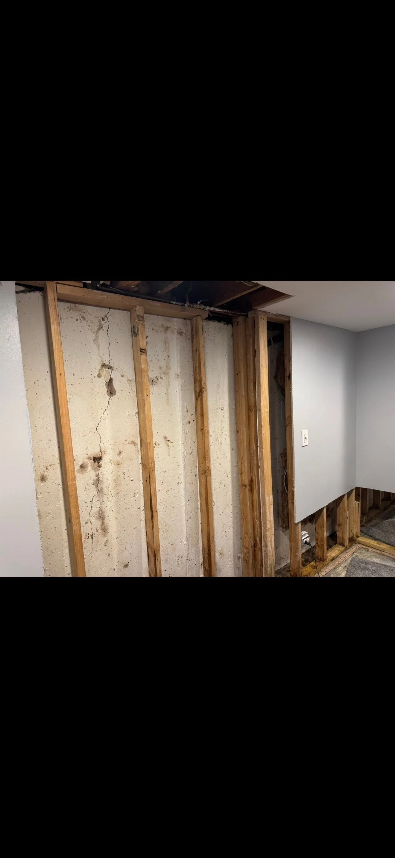 Interior wall demolition showing exposed wooden studs, cracked drywall, and some electrical wiring, with part of the wall covered by a painted drywall panel.
