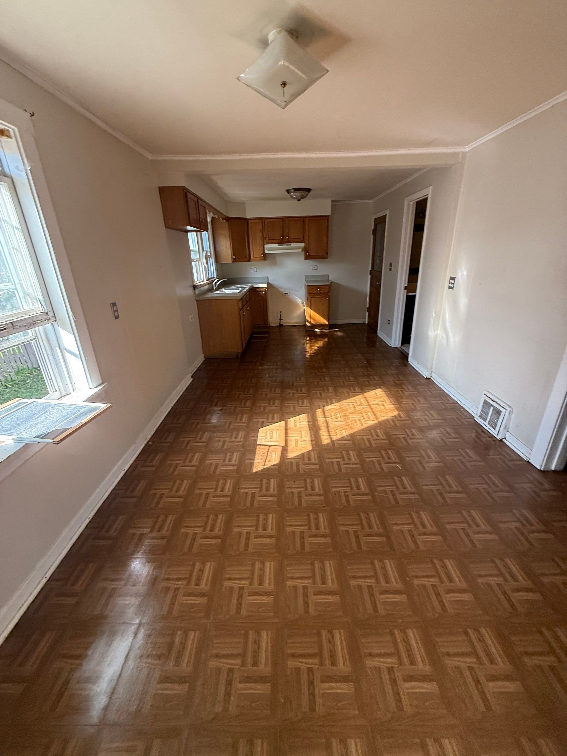 Empty living room with parquet flooring, natural light from a window, and an open kitchen area in the background.