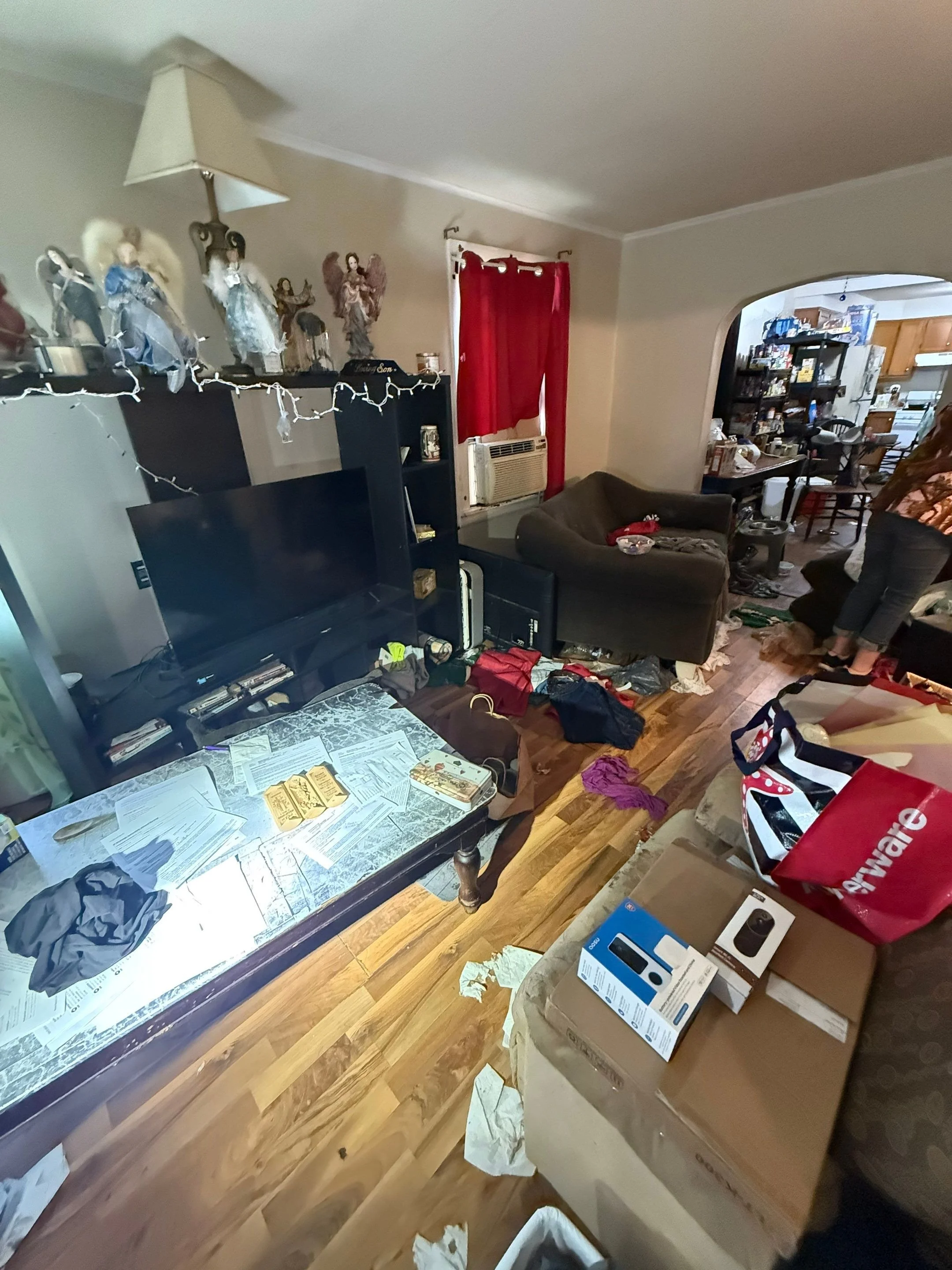Living room with clutter, papers on the coffee table, a large TV, shelves with figurines and decorations, a brown armchair, red curtains, and a person partially visible.