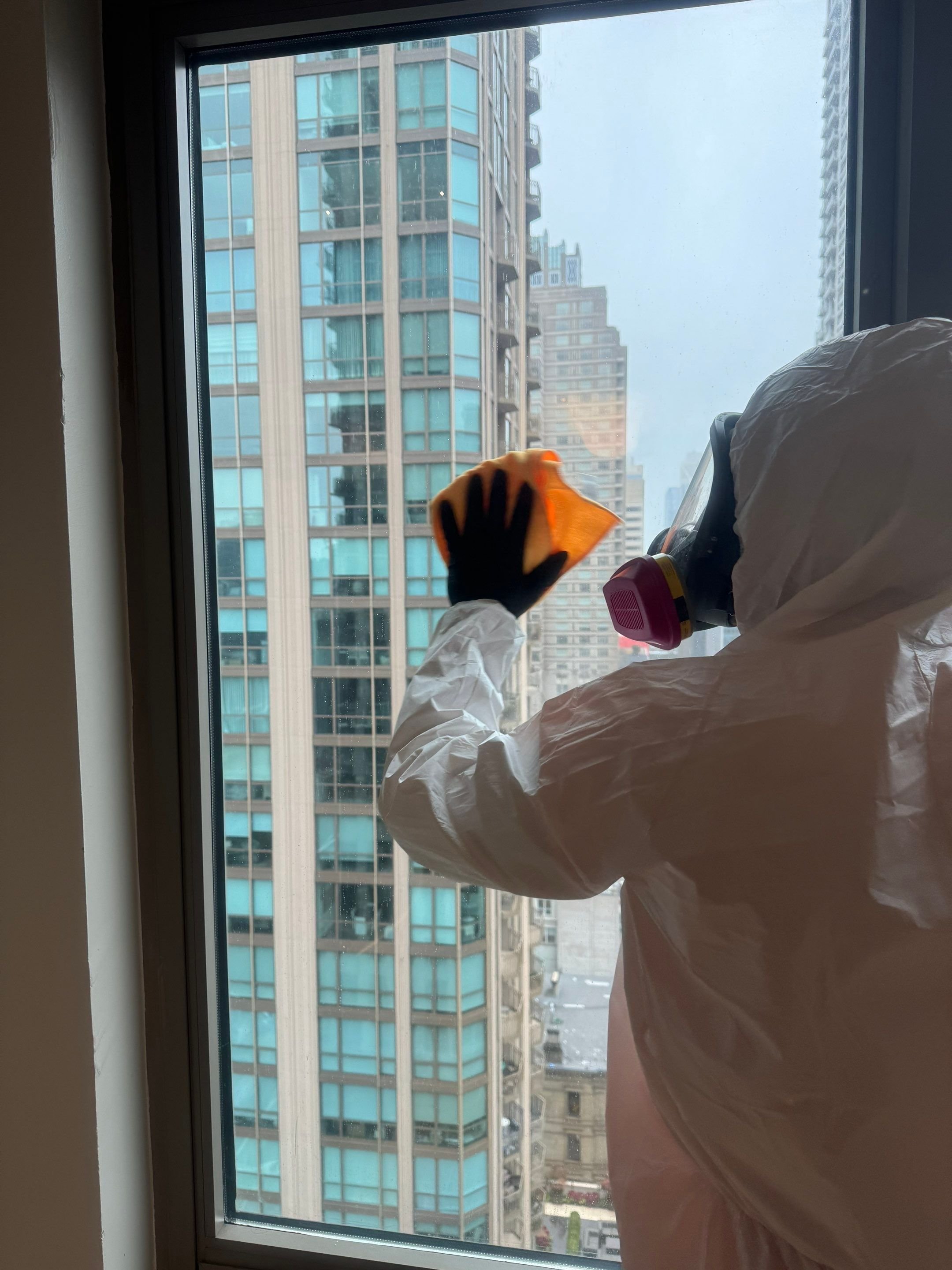 Person wearing protective gear, including a white suit and a respirator mask, cleaning a window in a high-rise building with a yellow cloth.