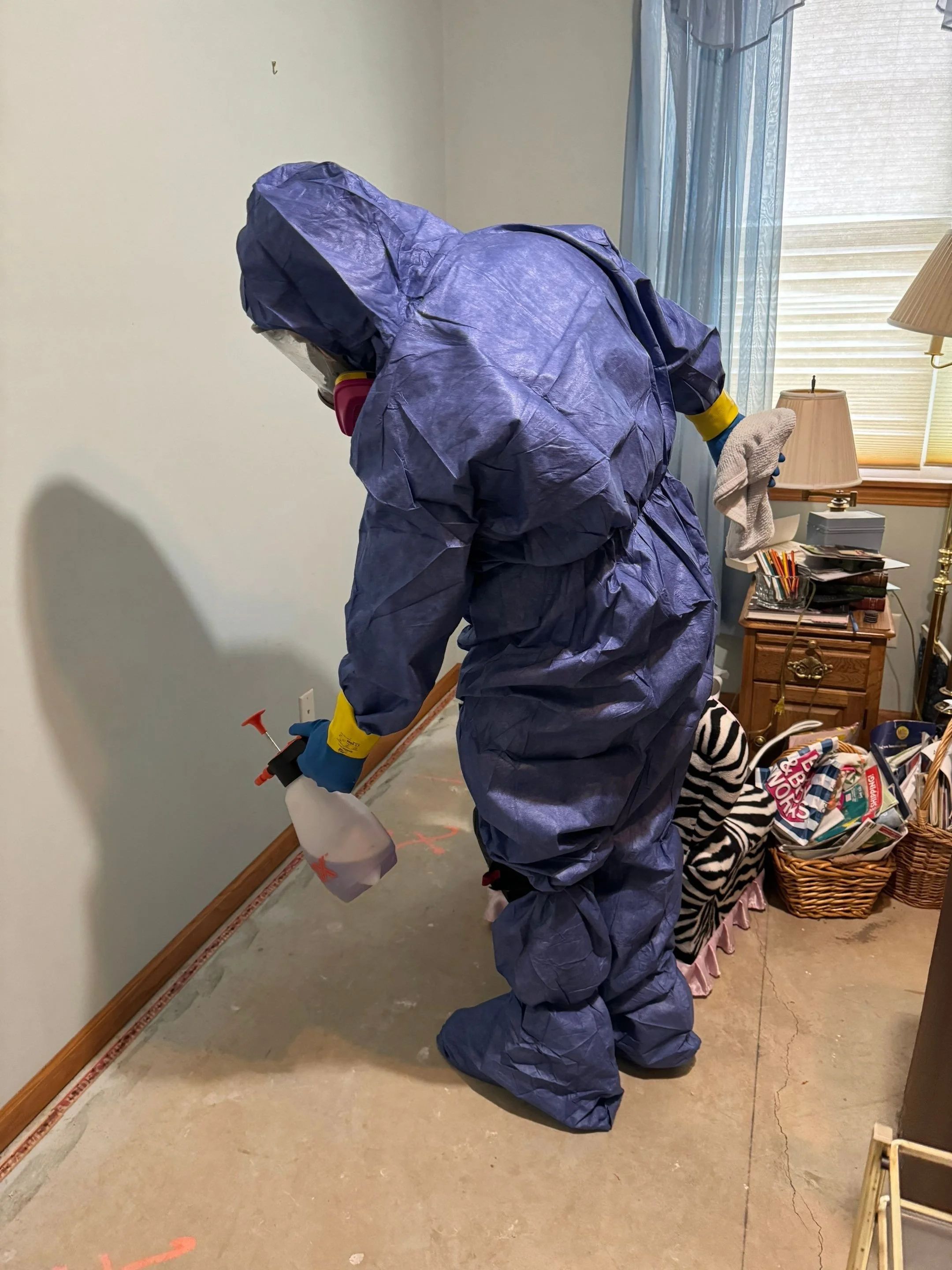 A person wearing a purple hazmat suit, gloves, and a respirator mask is spraying disinfectant in a room.