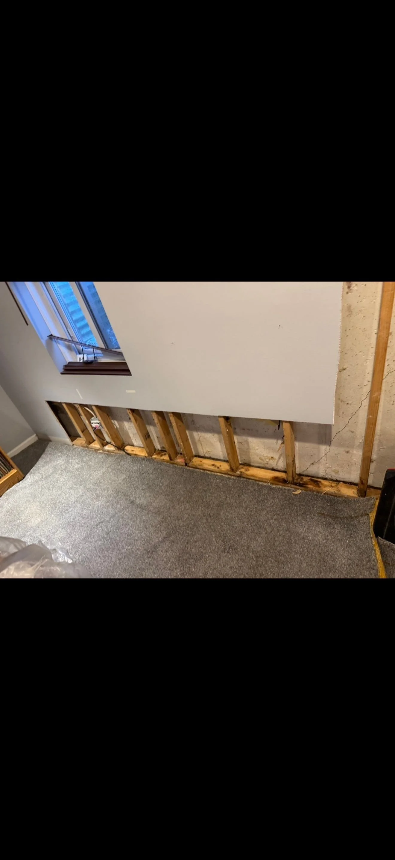 Partially removed wall exposing wooden studs and drywall, with carpeted floor and window at the top left corner.