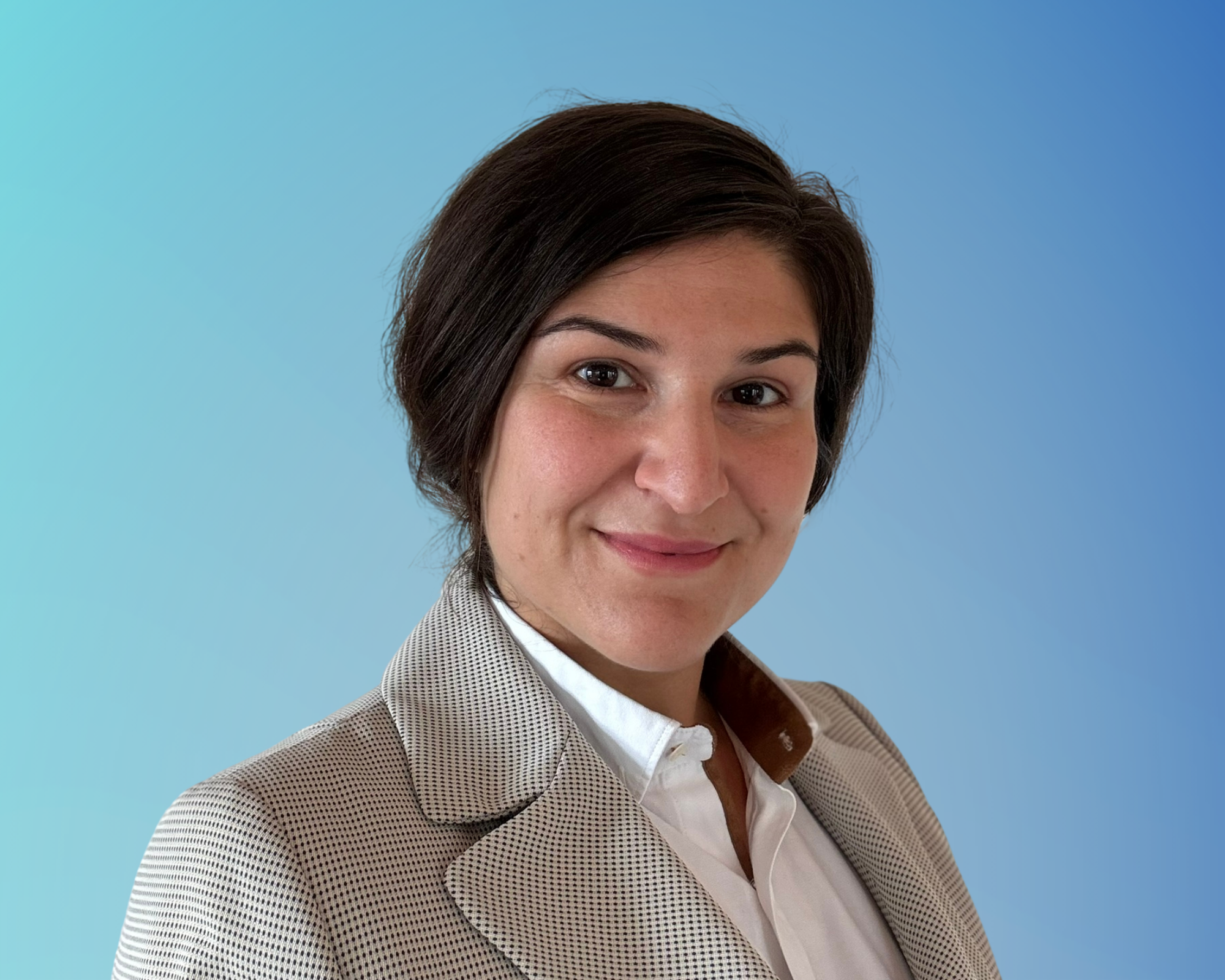 Porträt einer Frau mit kurzen dunklen Haaren, die lächelnd in die Kamera schaut, im Büro oder professionellen Umfeld, mit einem blauen Hintergrund.