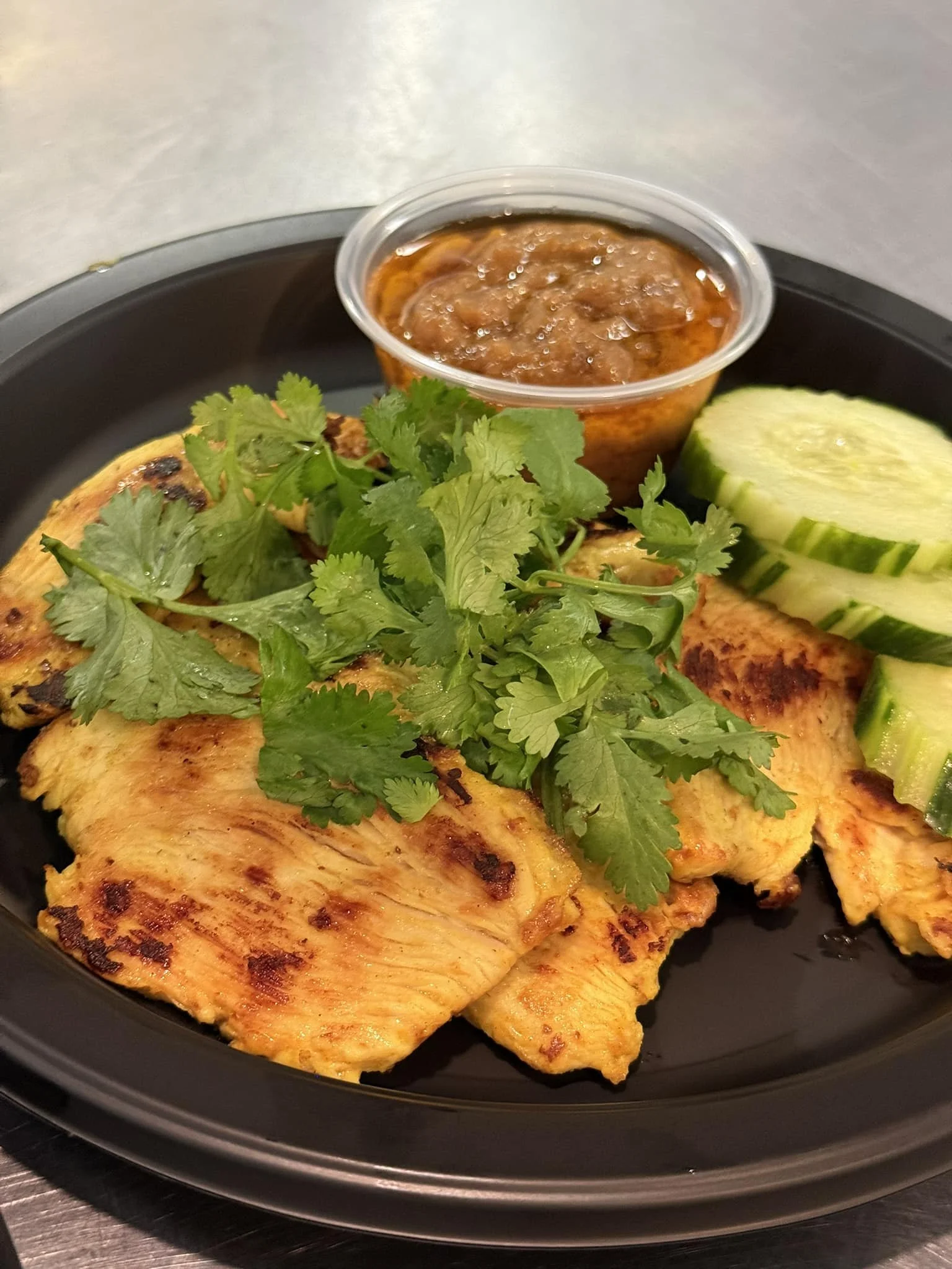 Grilled chicken with cilantro, sliced cucumbers, and a small container of chili sauce on a black plate.
