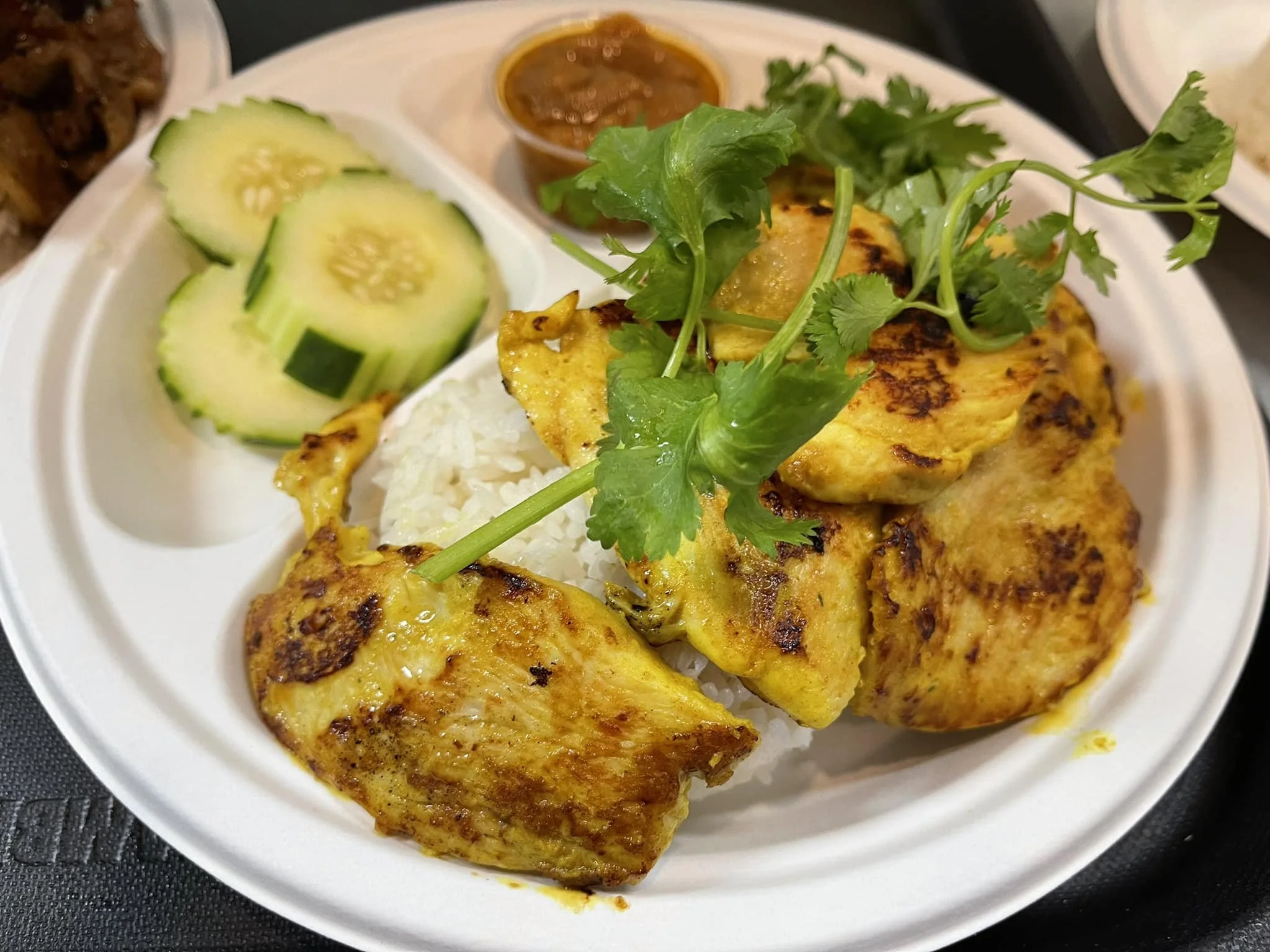 Plate of grilled chicken, white rice, cucumber slices, cilantro garnish, and a small container of sauce.