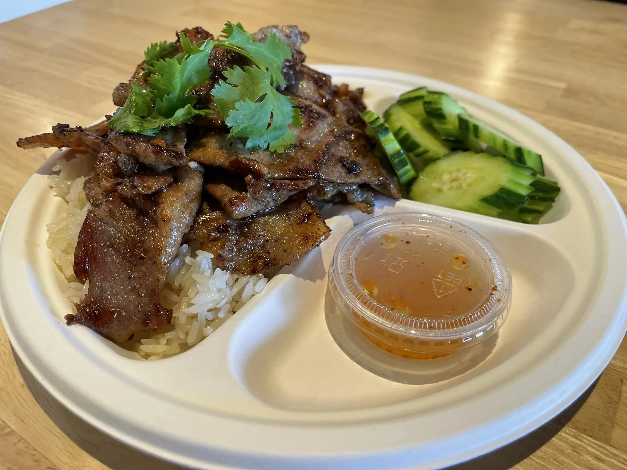 A takeout container with rice topped with grilled beef, garnished with cilantro, with sliced cucumbers and a small cup of dipping sauce on the side.
