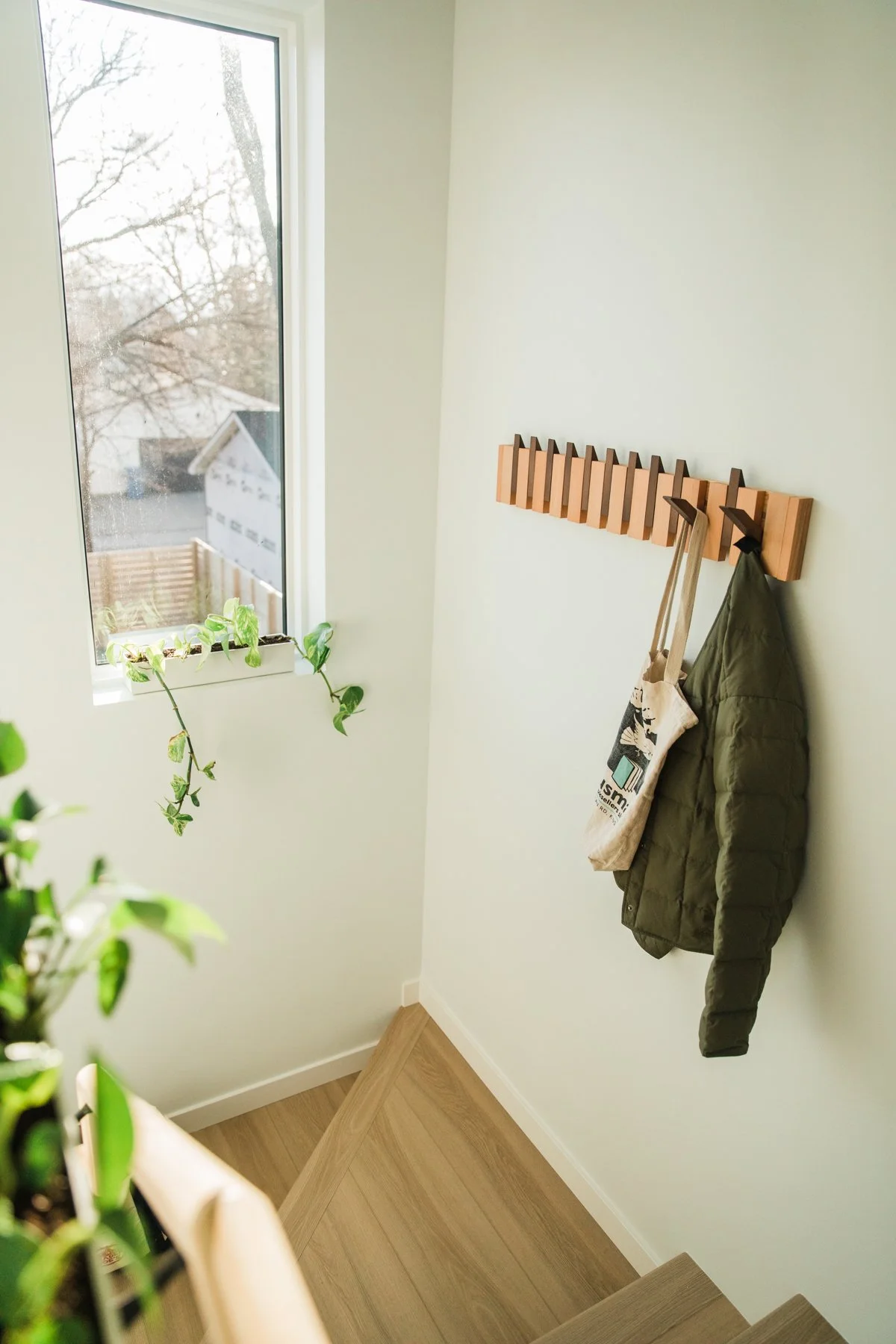 A corner of a room with a staircase, a large window, a wall-mounted wooden coat rack with hooks, a coat, and a tote bag hanging, and a green plant on the window sill.