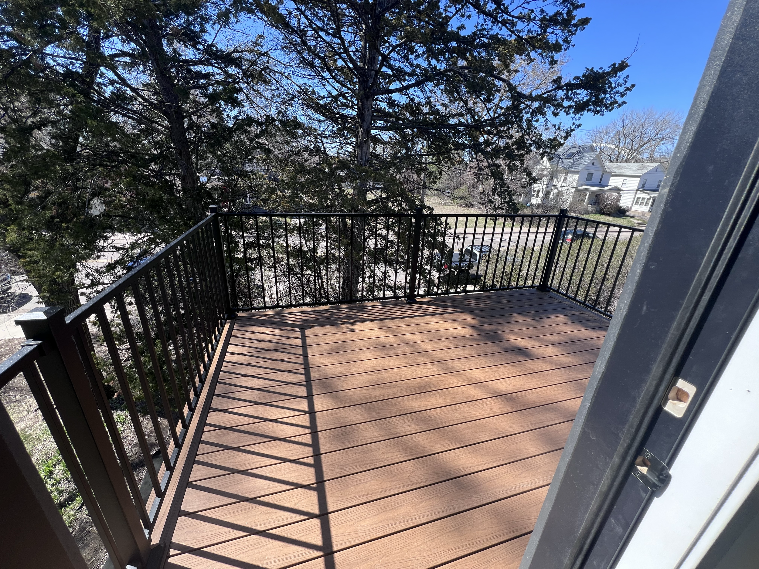 View from a balcony with a black metal railing, wooden floor, overlooking a residential neighborhood with white houses, trees, and parked cars on a sunny day.