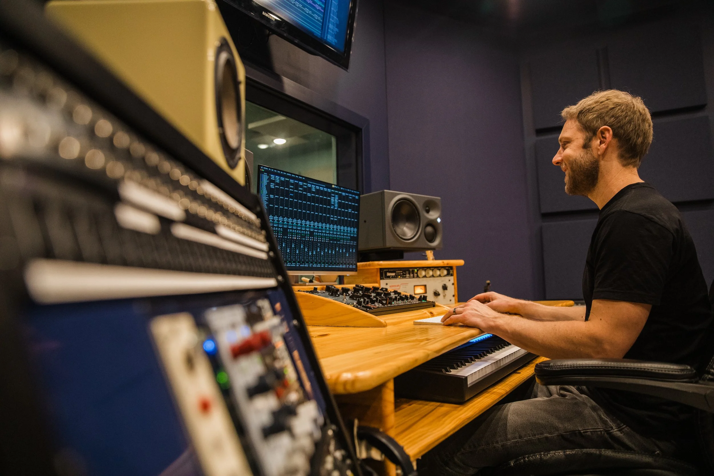 A man sitting at a keyboard in a music studio, smiling while looking at computer screens, with audio equipment and sound monitors around him.