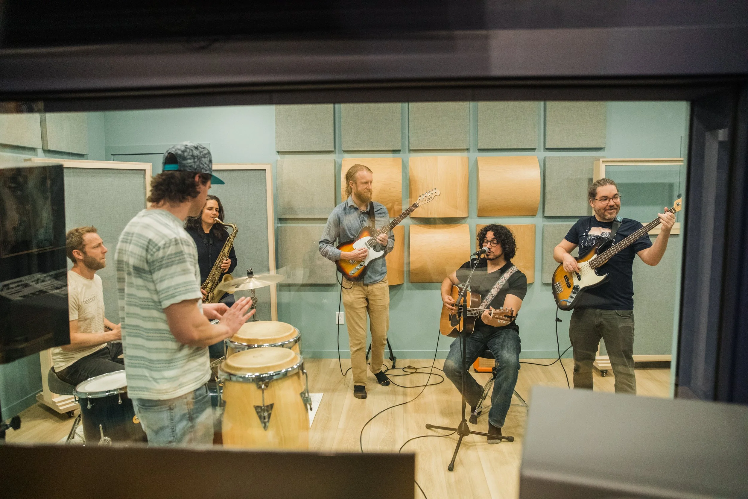 A group of musicians practicing in a recording studio, playing various instruments including guitar, bass, drums, and saxophone, with acoustic paneling on the walls.