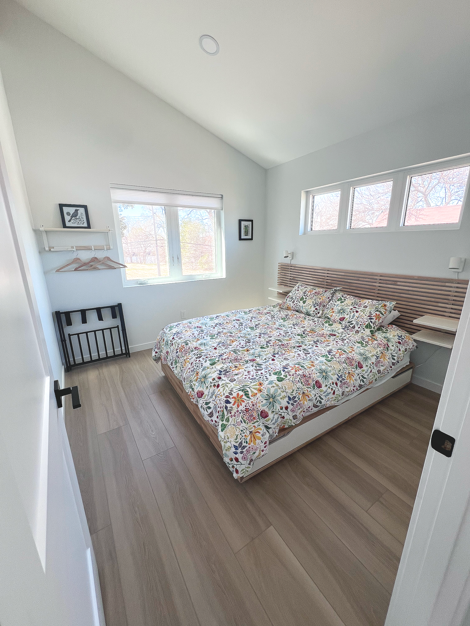 A bedroom with a bed covered with a floral bedspread, a wooden headboard, and a large window showing an outdoor scene with trees and a blue sky.