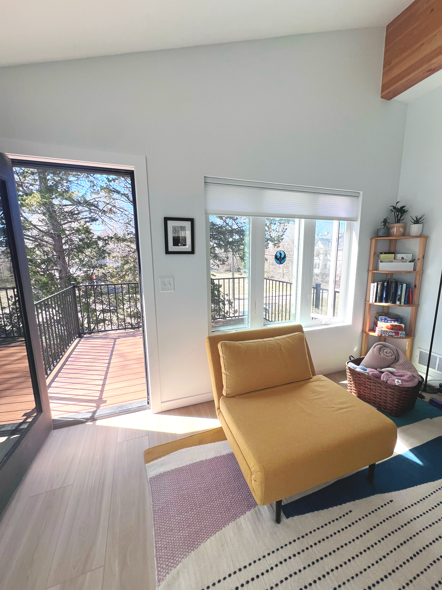 A living room with a yellow chaise lounge, a patterned rug, a wooden bookshelf, a basket with blankets, and a sliding glass door opening to a balcony with trees outside.