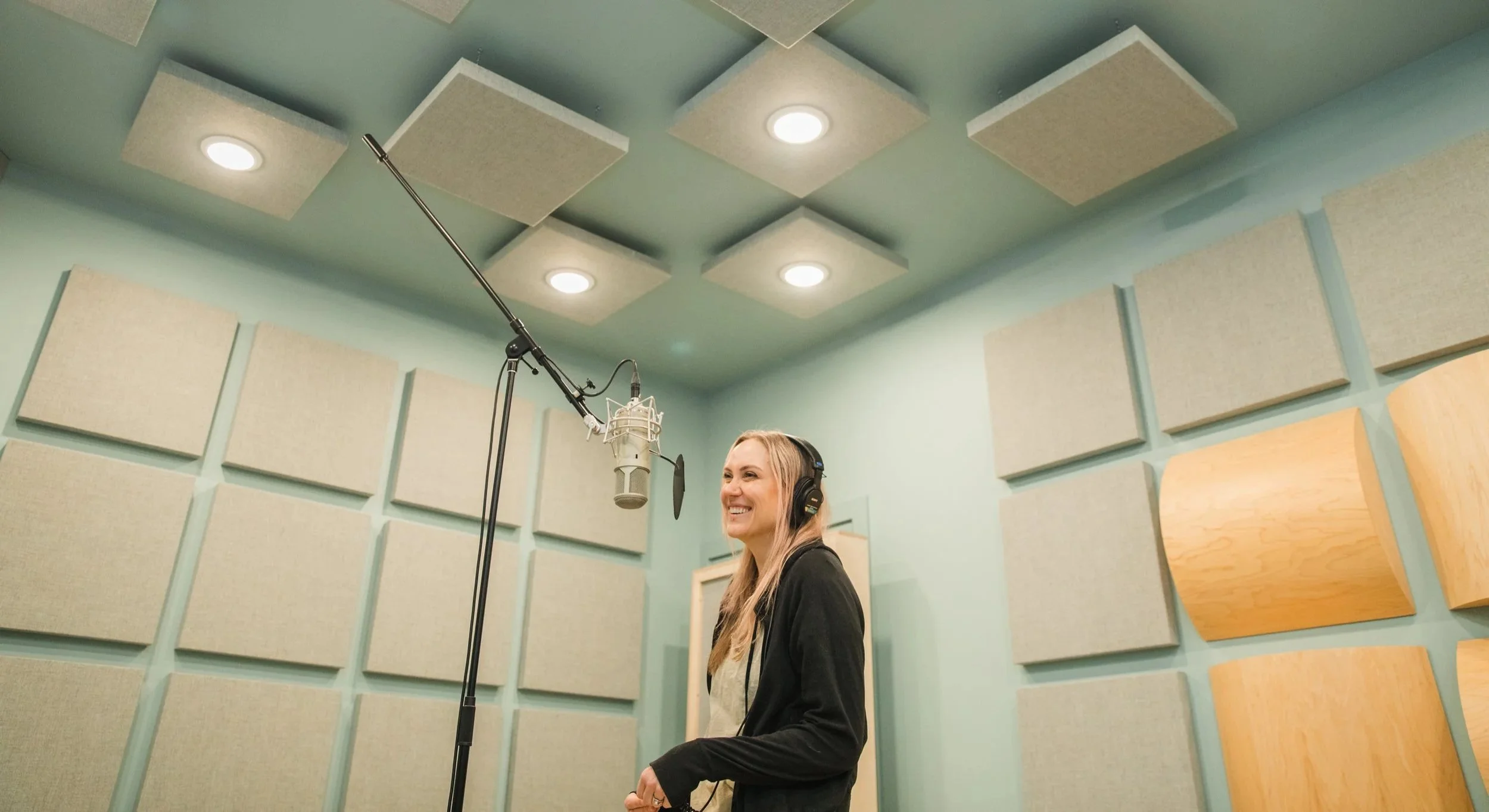 A woman with long blonde hair, wearing a black blazer, smiling while standing in a recording studio with beige acoustic panels on light blue walls, wearing headphones, and positioned in front of a professional microphone.