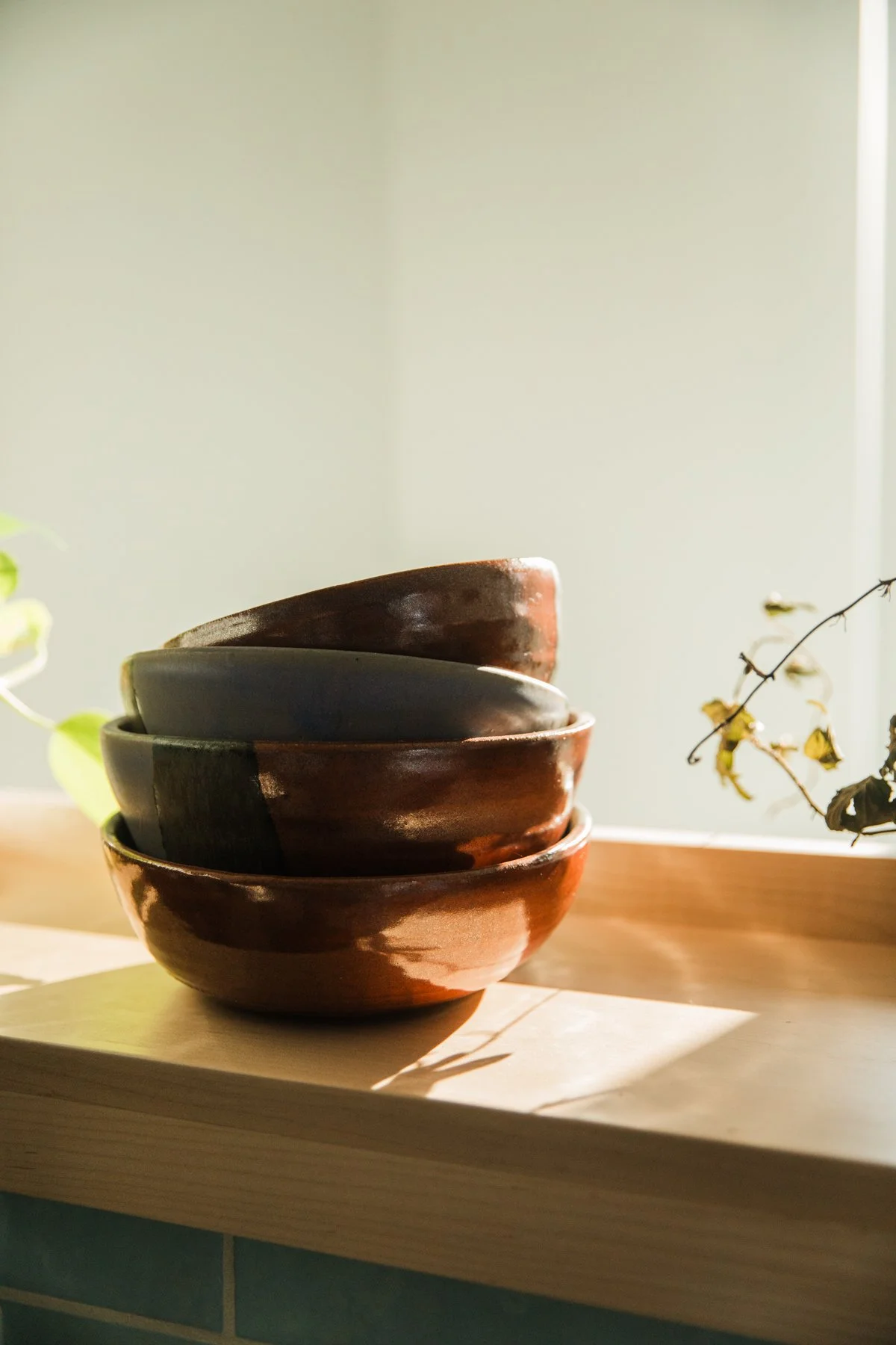 A stack of five ceramic bowls with different shades of brown and gray, placed on a wooden surface near a window with sunlight.