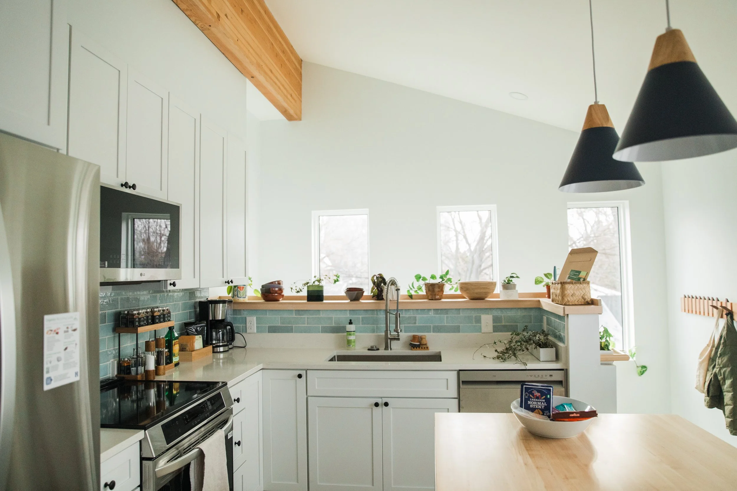 A modern white kitchen with a green tiled backsplash, white cabinets, a microwave, and a counter with plants and bowls, under a sloped ceiling with wooden beams and large windows.