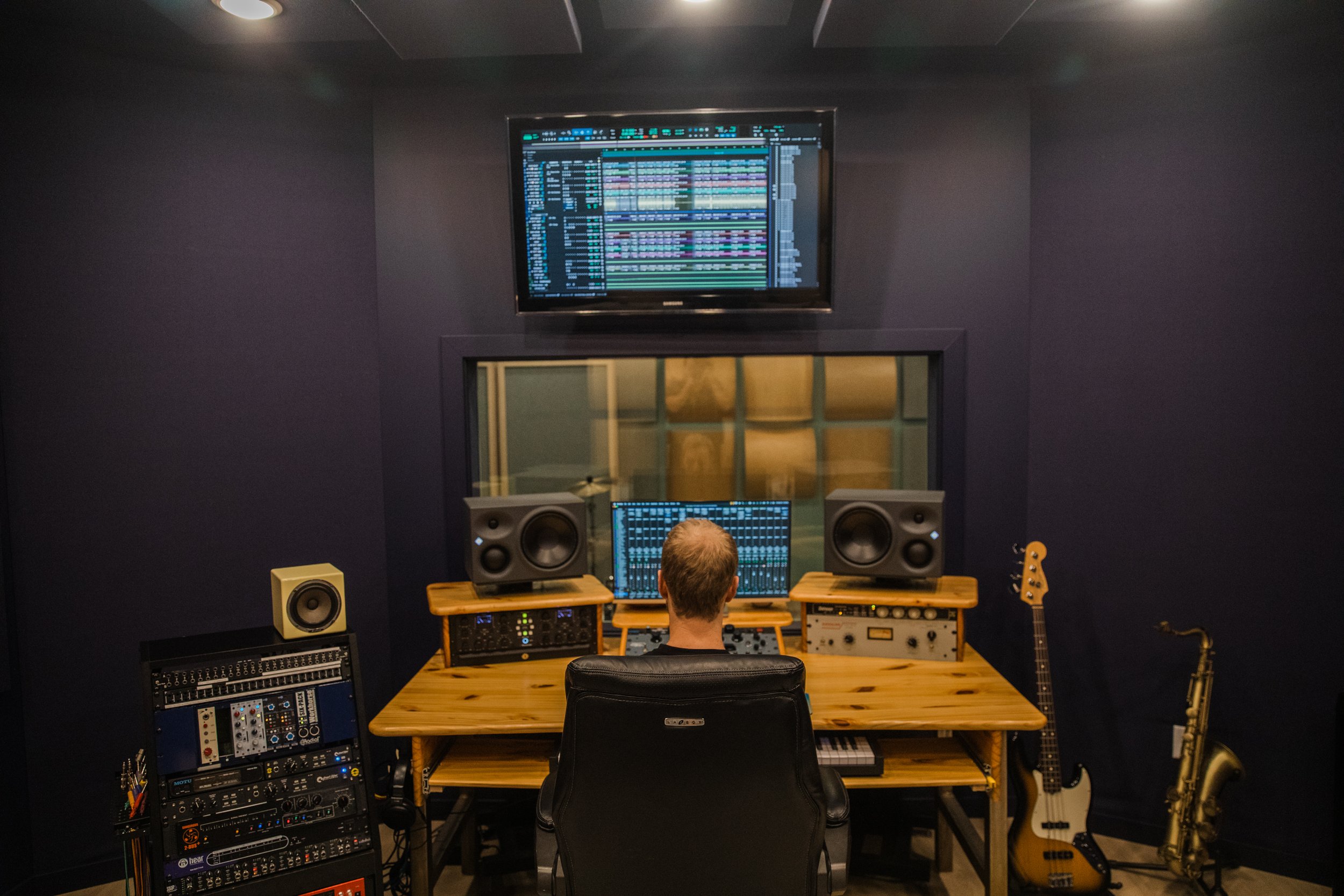 A person sitting in a music production studio, facing a computer screen displaying digital audio workstation software. Surrounding the person are studio monitors, audio equipment, and musical instruments including a guitar.