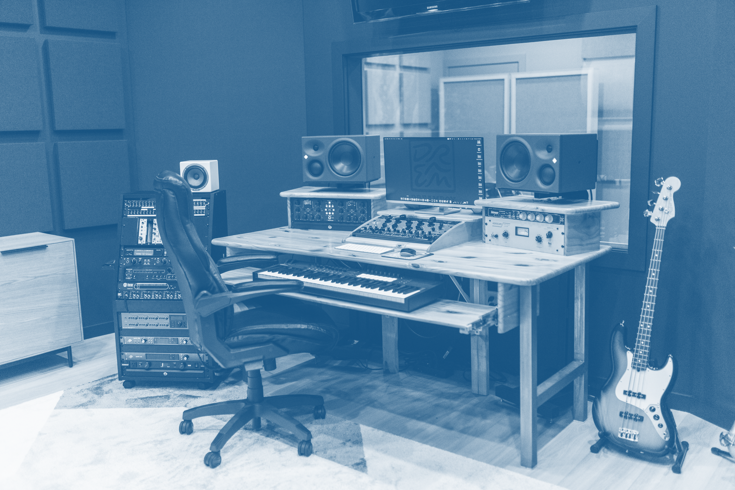 A music recording studio control room with a desk holding speakers, audio equipment, a computer monitor, and a keyboard, with a guitar leaning against the desk.