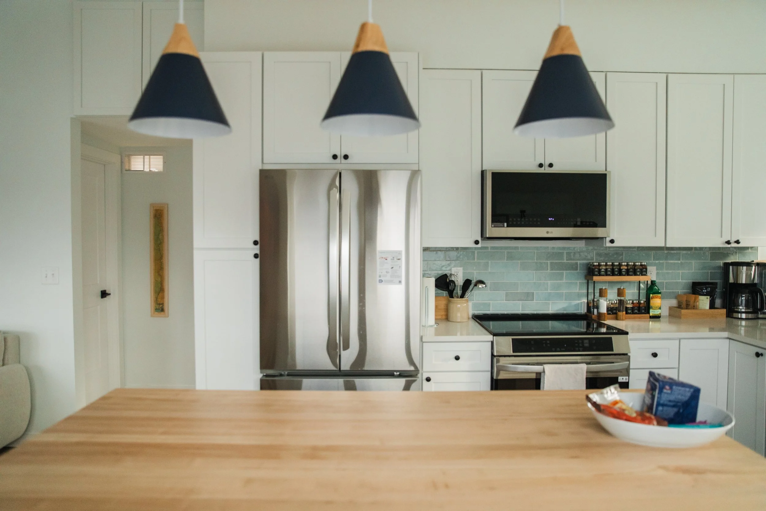 A kitchen with white cabinets, stainless steel refrigerator, black microwave, and stovetop. A wooden table in the foreground has a bowl of snacks on it. Three black and wood cone-shaped pendant lights hang above the table.