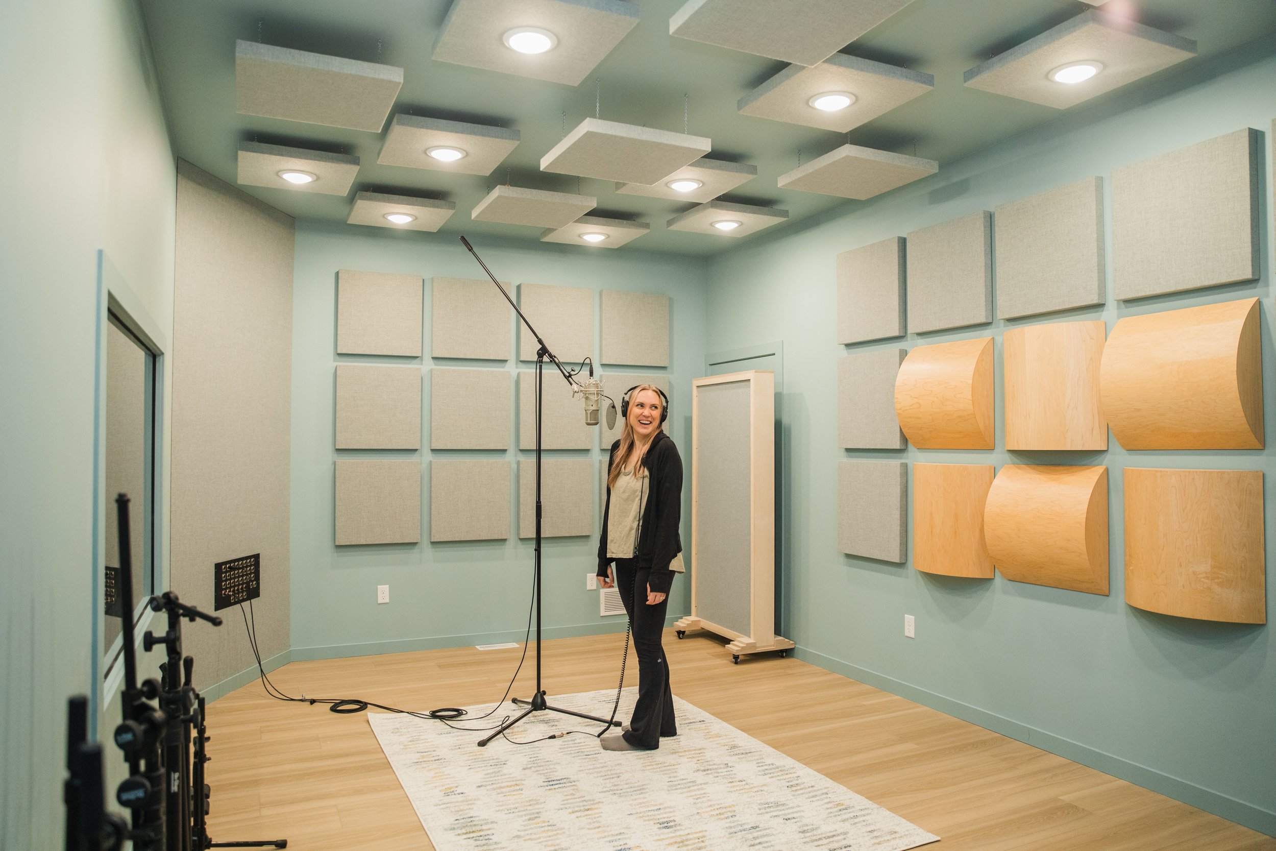A woman wearing headphones and a black jacket standing in a recording studio with soundproofing panels on the walls, standing near a microphone stand on a white rug.