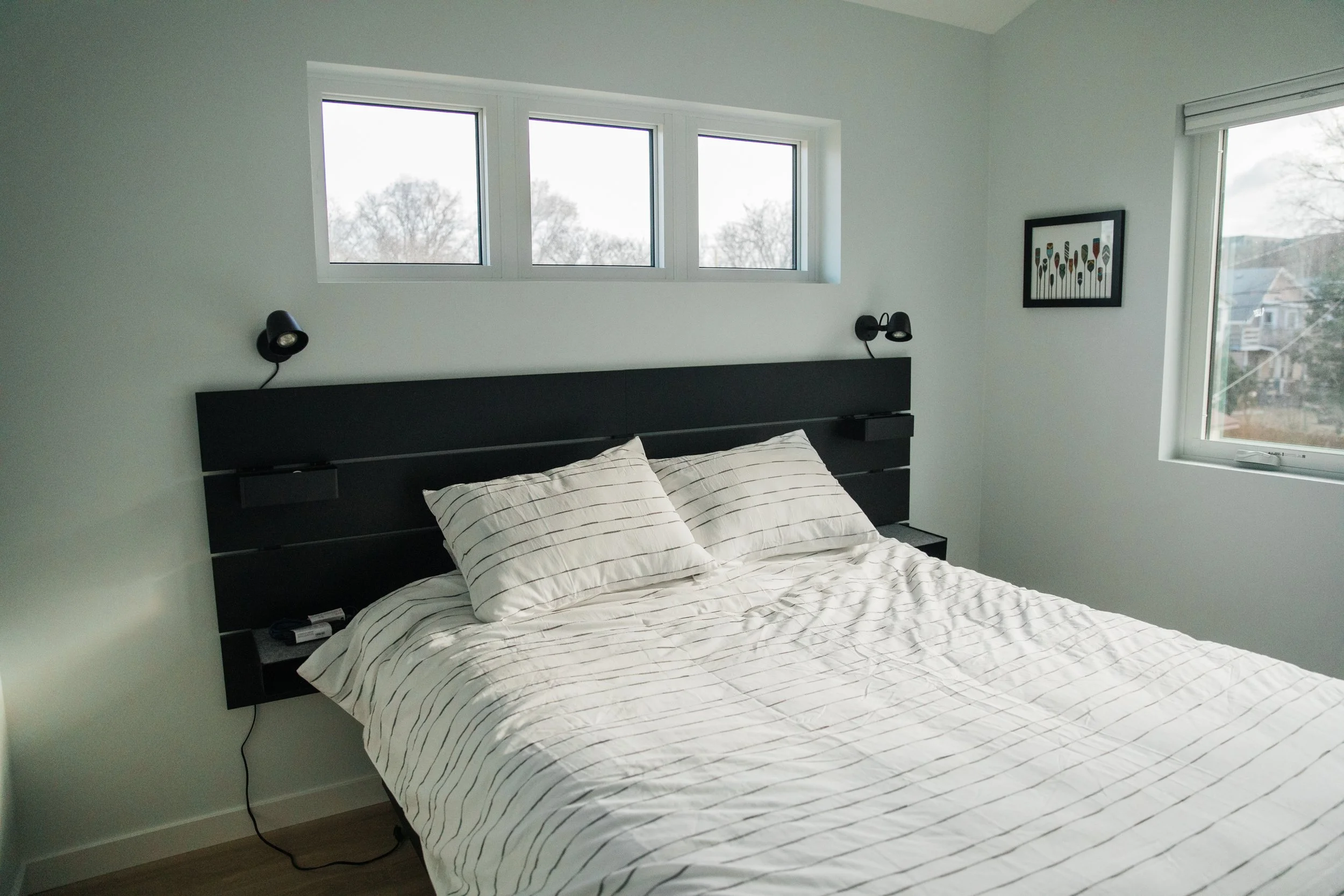 A bedroom with a bed, striped bedding, two pillows, wall-mounted lamps, a window with three panes showing trees outside, a framed artwork, and another window to the right with a view of neighboring houses and trees.