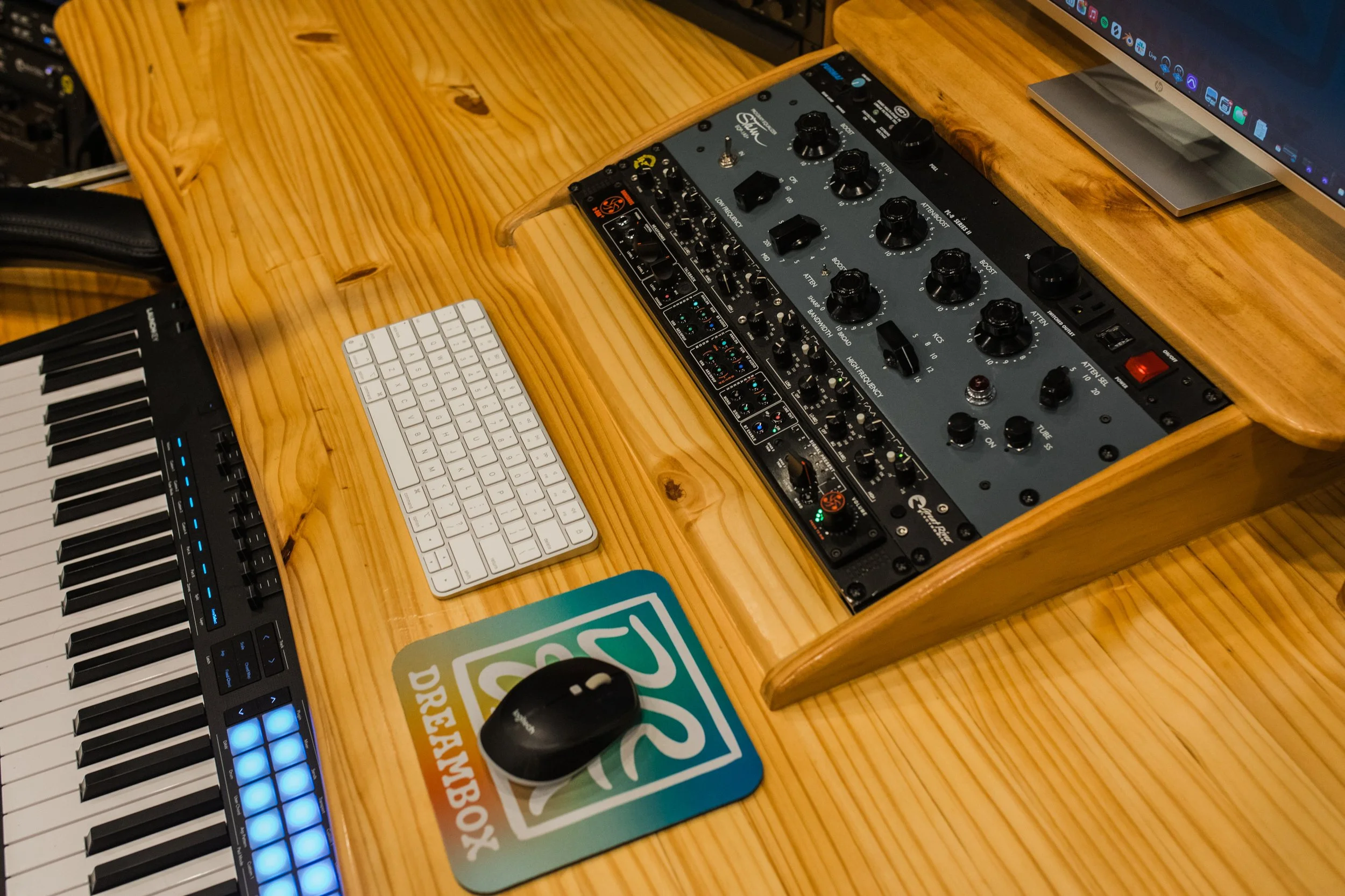 A wooden desk with a computer monitor, a MIDI keyboard, a small audio mixer, a white wireless keyboard, a black computer mouse, and a colorful sticker or coaster.