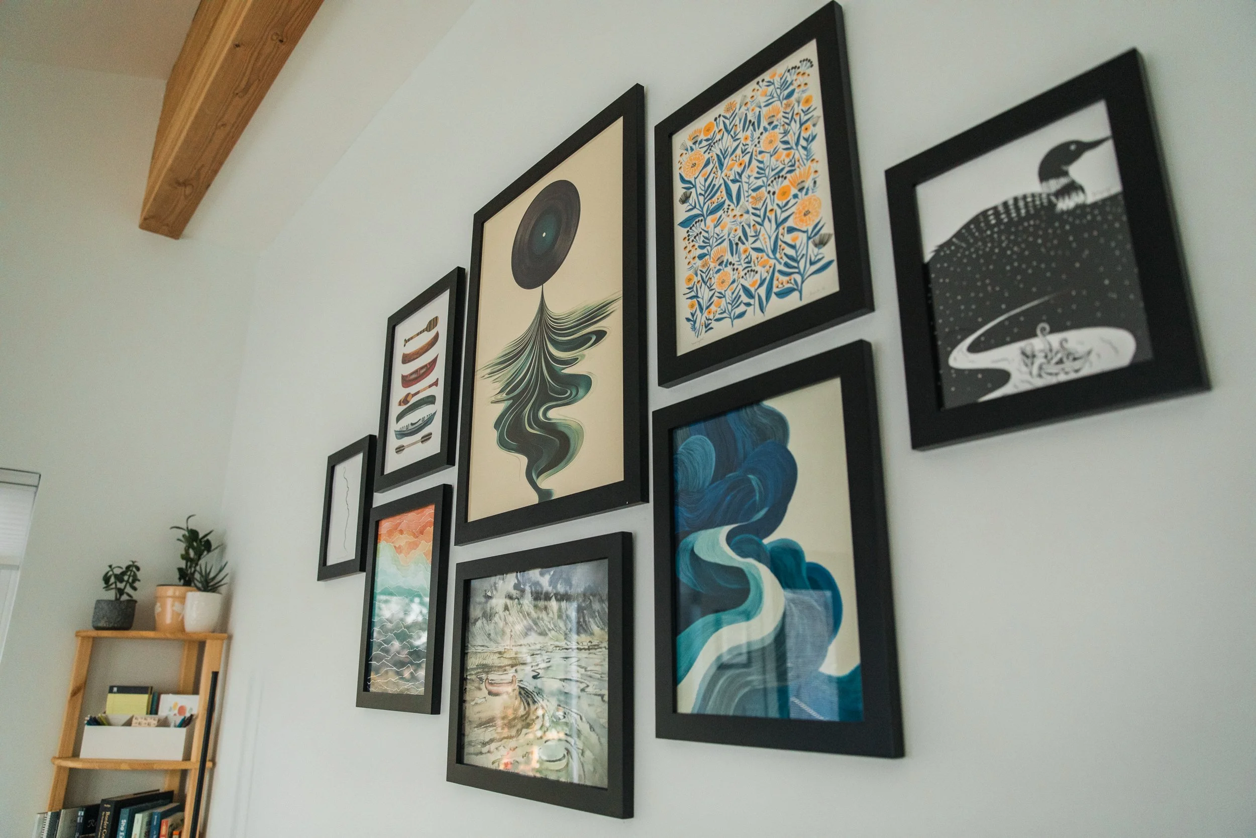 A gallery wall with multiple framed artworks, including abstract, nature scenes, and graphic designs, in black frames. A wooden shelf with potted plants and books is visible below, and a wooden beam runs along the ceiling.