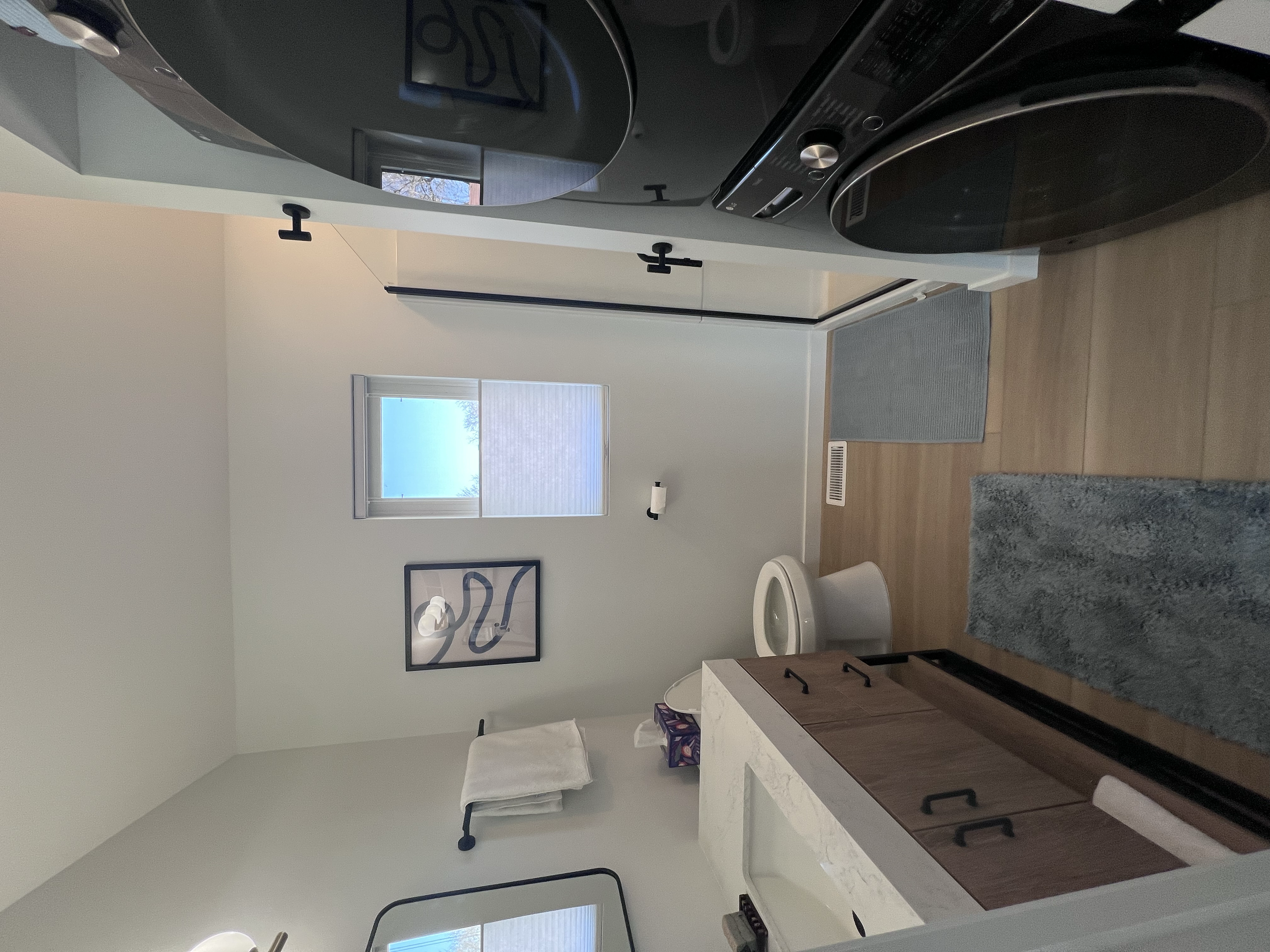 A bathroom with a white toilet, a stone vanity with bronze cabinets, a mirror, and two framed abstract art pieces on a light wall. There is a window allowing natural light and a small rug on the wooden floor.