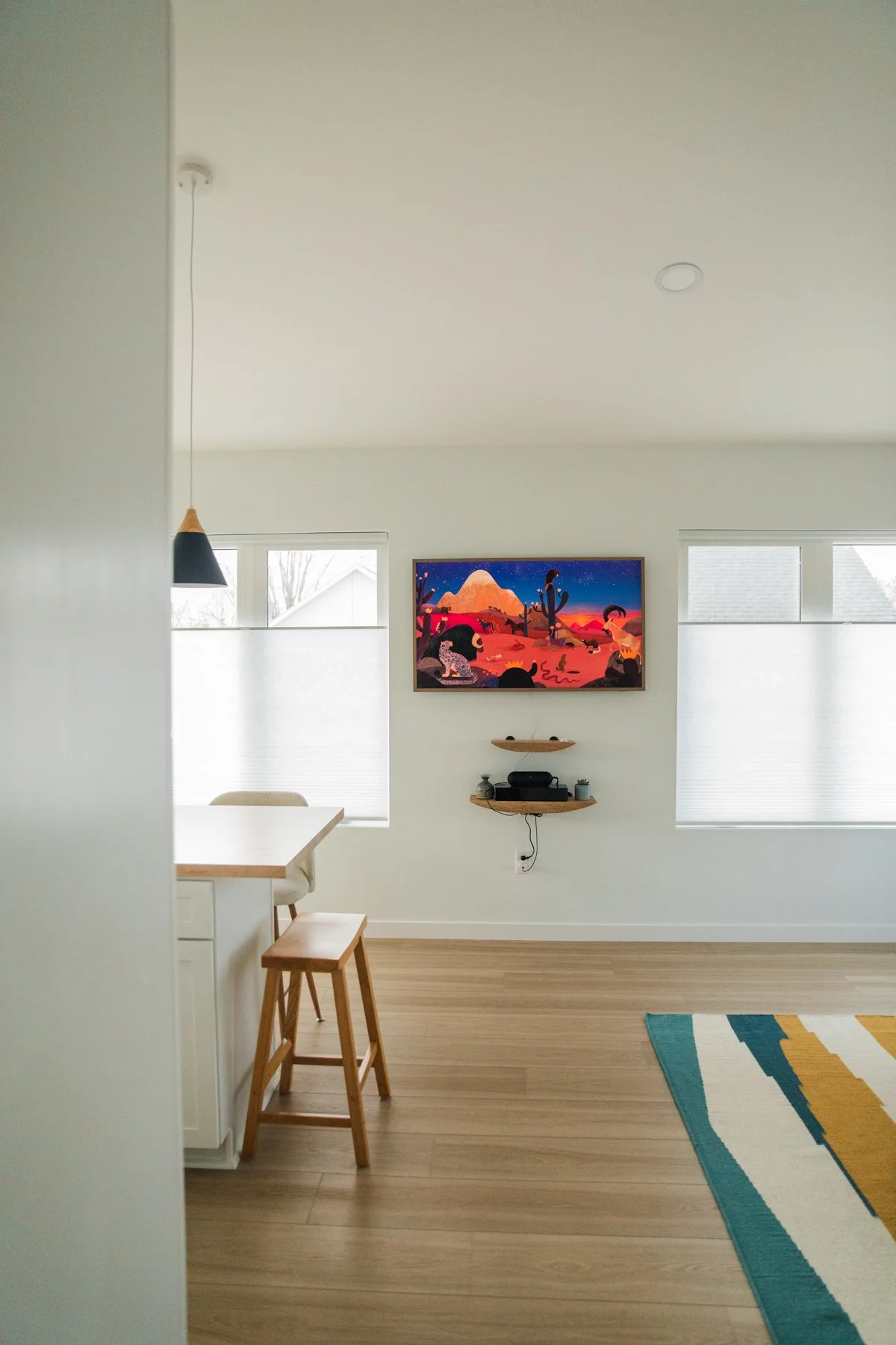 A minimalist living room with light wood flooring, a large colorful landscape painting on the wall, a floating shelf below the painting with electronic devices and small potted plants, two windows with blinds, a dining counter with a stool, and a par