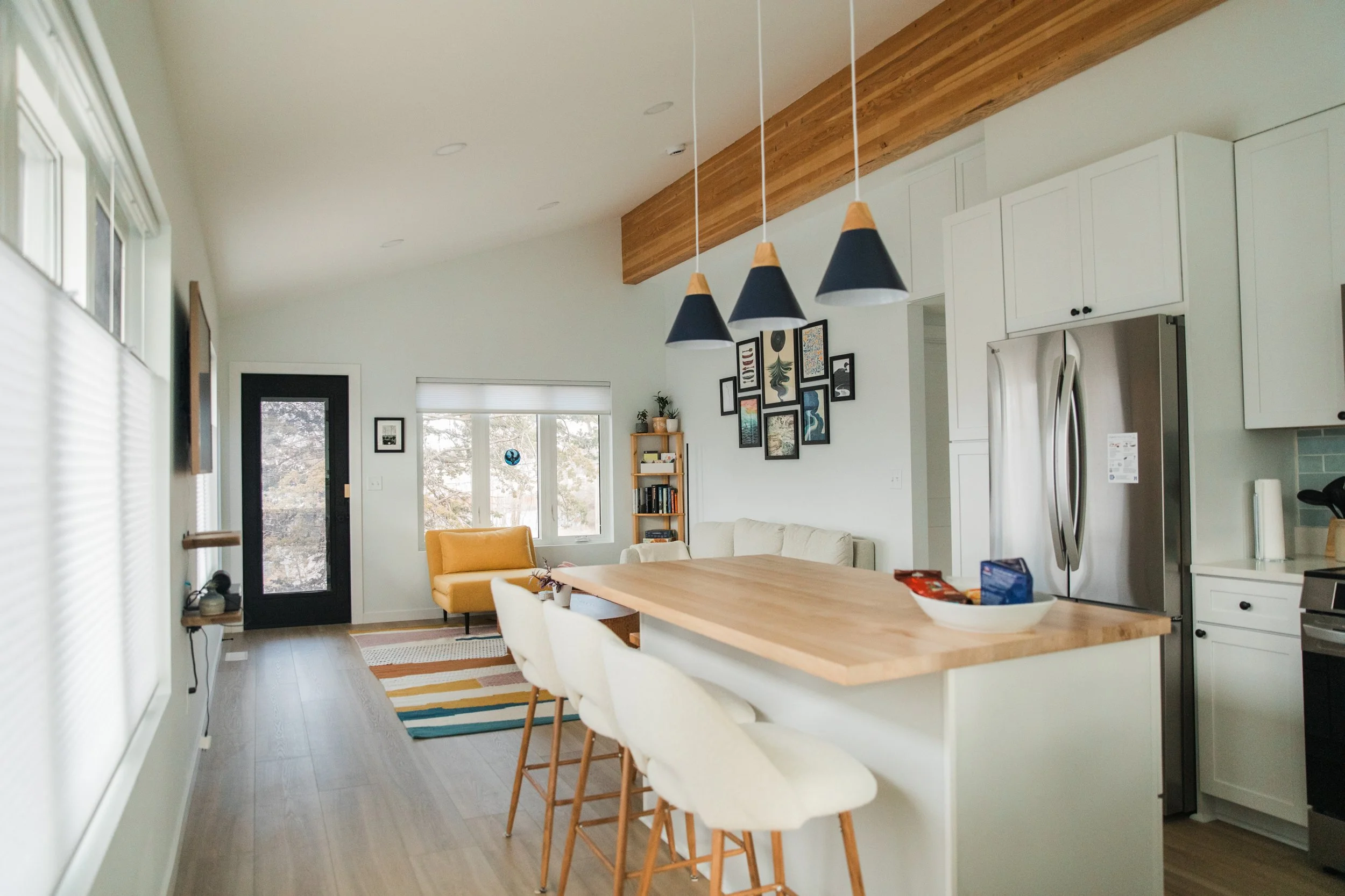 Open-concept living space with wooden and white accents, featuring a dining area with white chairs, a yellow armchair, a living room with wall art, and kitchen appliances including a stainless steel refrigerator.
