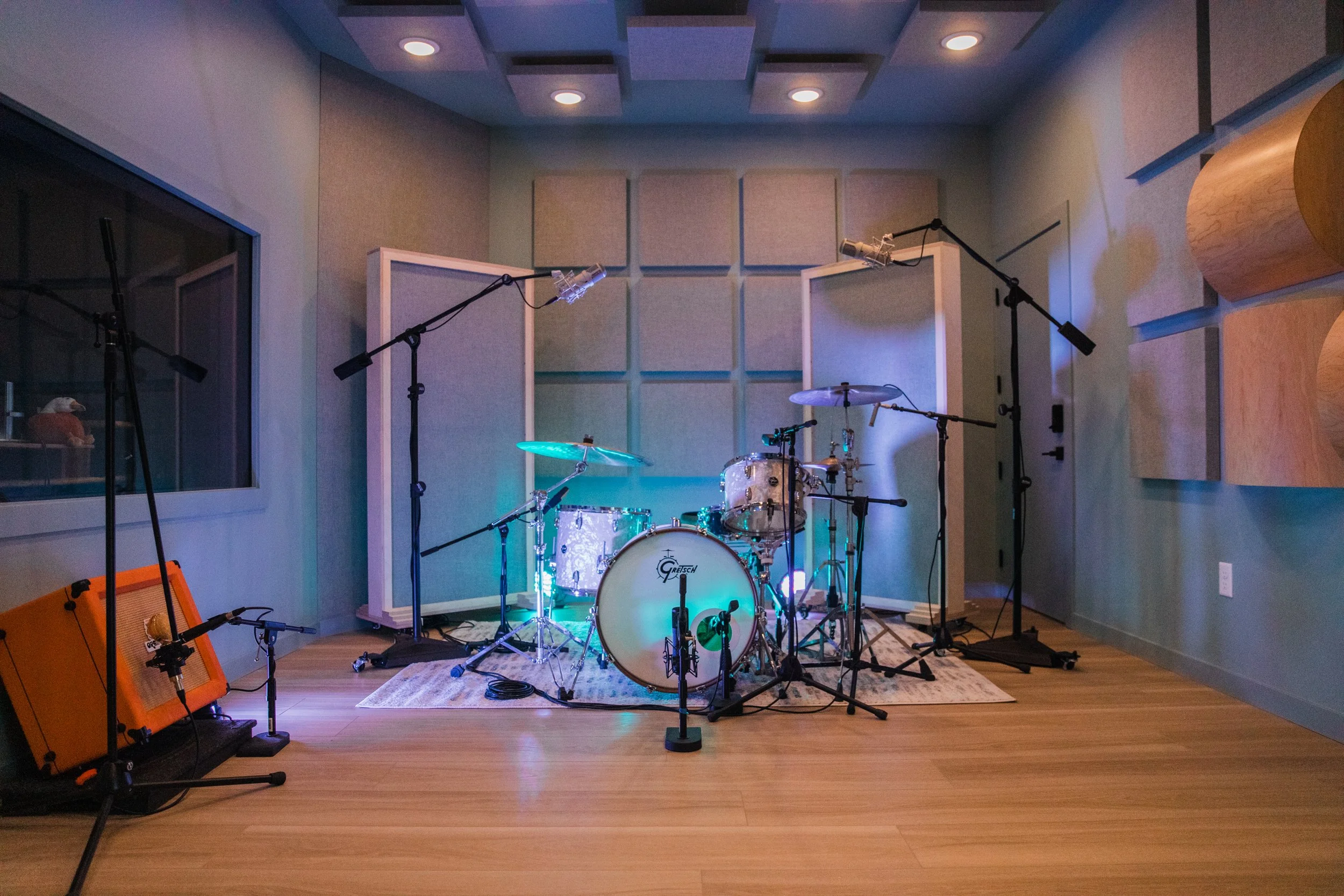 A music recording studio with a drum set on a rug, microphones, an orange amplifier, and soundproofing panels on the walls.