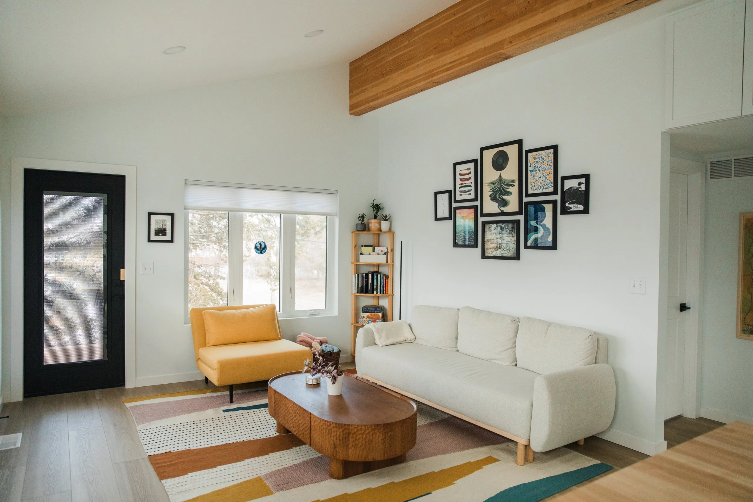 Living room with white sofa, yellow armchair, wooden coffee table, wall art, window with blinds, small bookshelf, and a door with a screen.