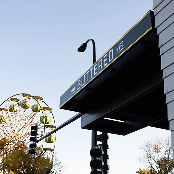 Street view featuring a black sign with yellow and white text that reads "The Buttered Tin," a Ferris wheel in the background, a streetlight, and traffic lights.