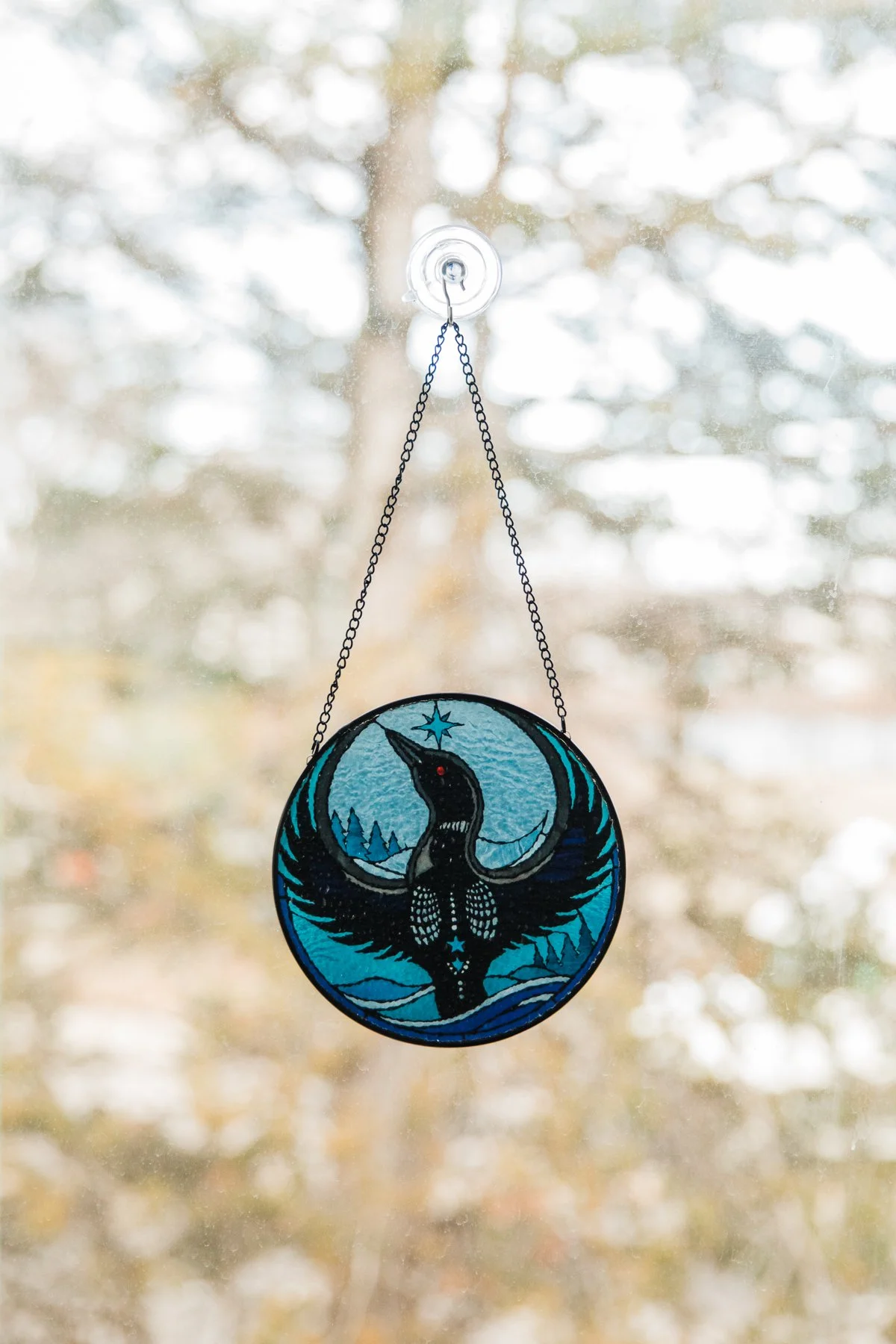 A stained glass ornament featuring a dark bird with outstretched wings, a long curved beak, and a star above its head, hanging from a chain on a window with a blurry outdoor view in the background.