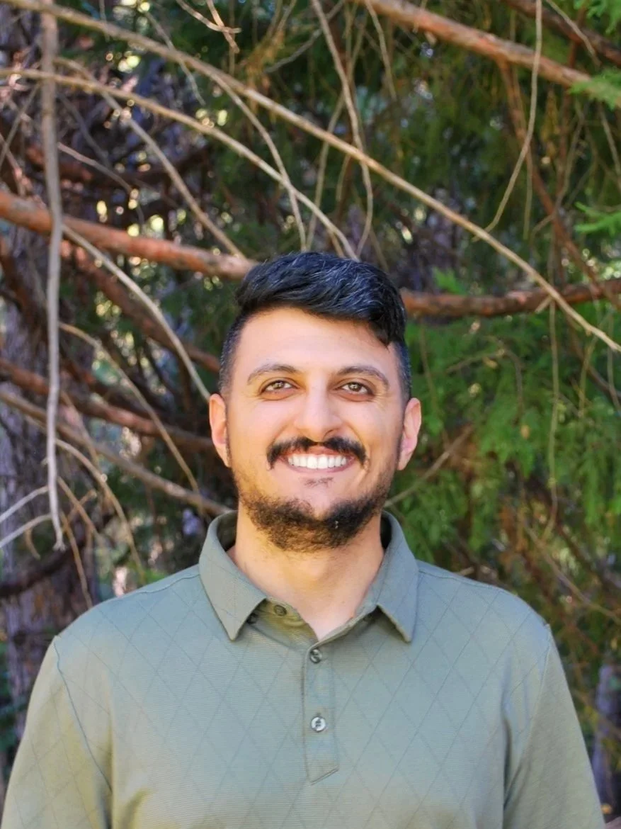 A smiling man with dark hair and a beard, wearing a light green collared shirt, standing outdoors in front of a background of trees.