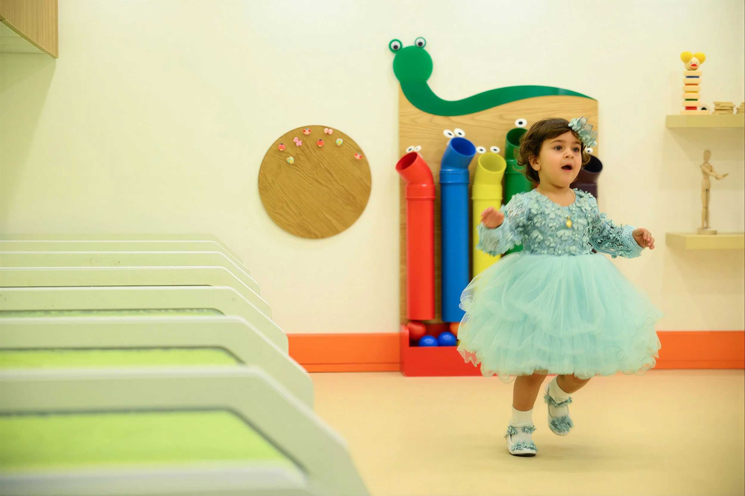 A young girl wearing a blue tutu and floral headband running in a colorful playroom, with water pipe and educational toys in the background.