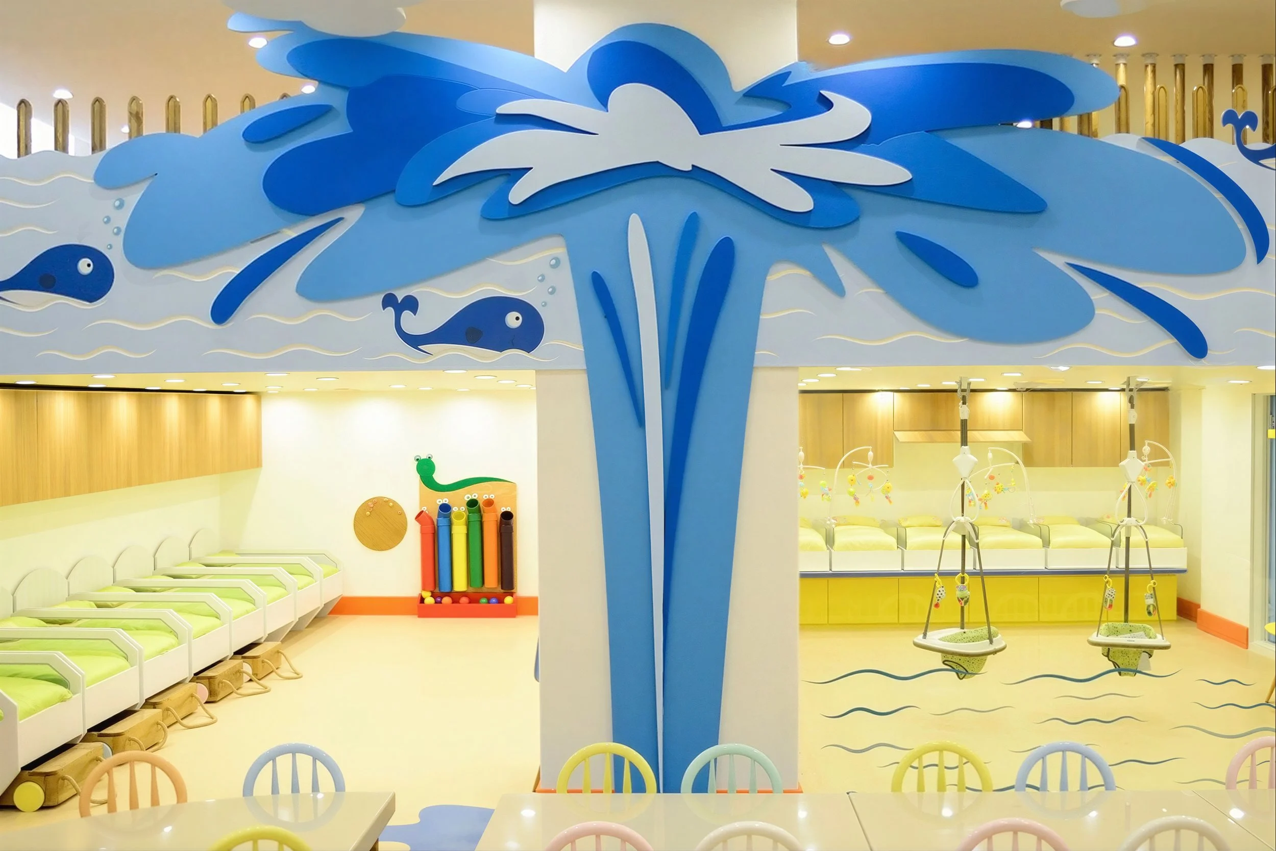 A colorful children's indoor play area decorated with a blue and white whale-themed structure. The room features small beds with green mattresses, a wall-mounted wooden toy with colorful tubes, and swings with blue and yellow seats. The background has warm wooden panels and soft lighting.