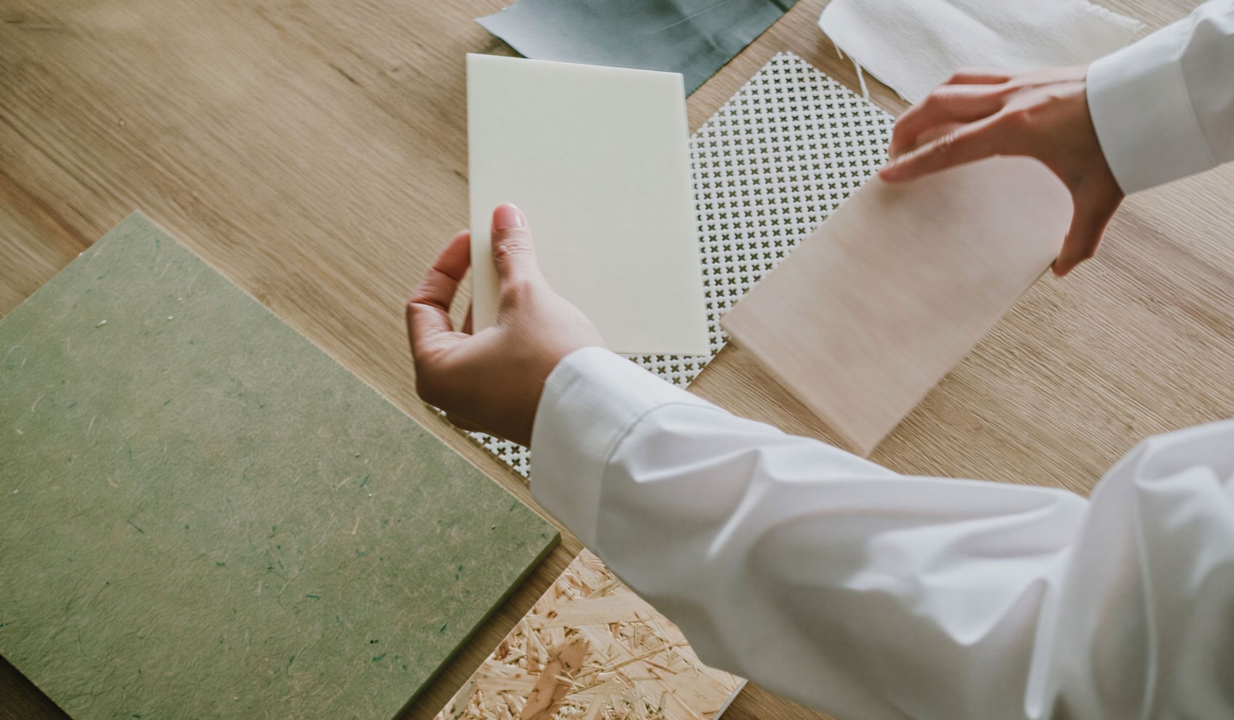 Person in a white coat flipping through samples of different materials on a wooden table, including fabric and wood samples.