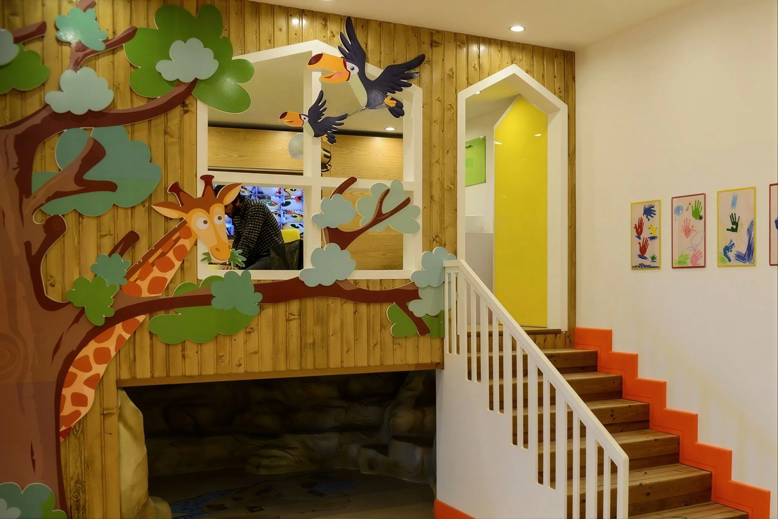 Children's playground with jungle animal theme, featuring tree and animal cutouts, including giraffe and toucan, with colorful artwork on the walls and a staircase with orange accents.