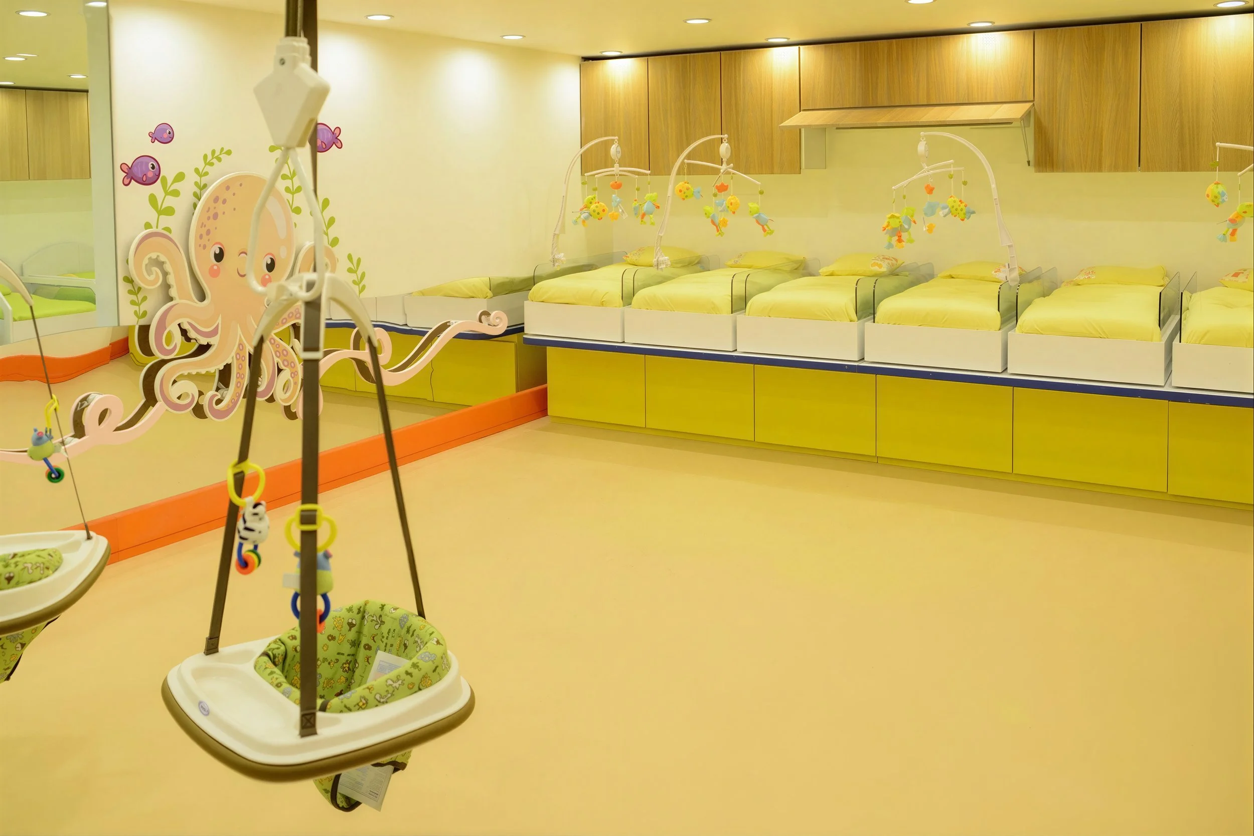A children's hospital or nursery play area with multiple cribs and hanging mobiles, decorated with a large pink octopus cartoon on the wall, painted fish, plants, and brightly colored bedding and toys.