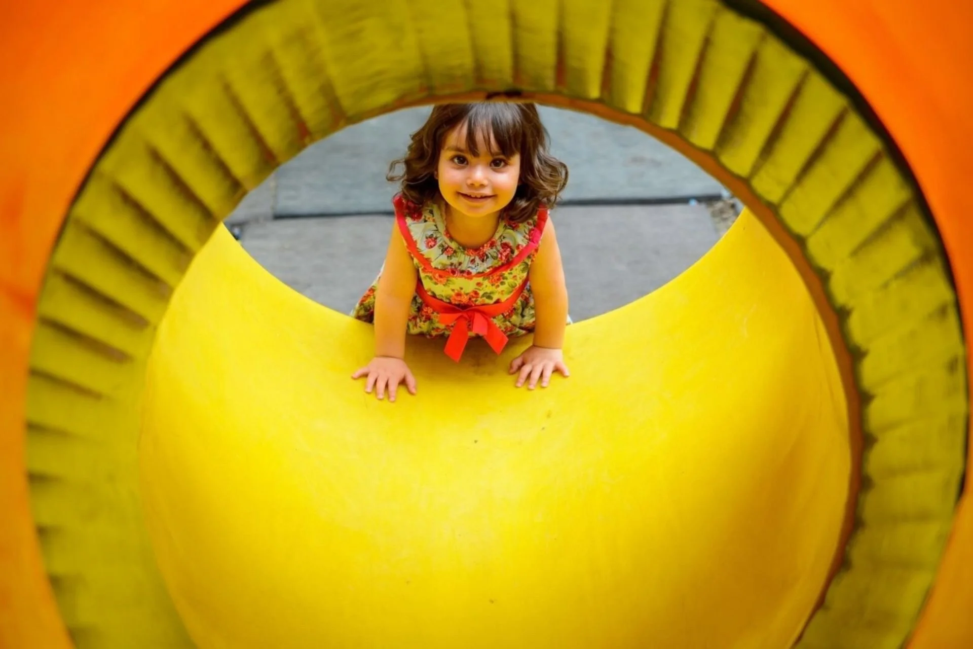 A young girl crawling through a yellow playground tunnel with orange edges, smiling and looking at the camera.