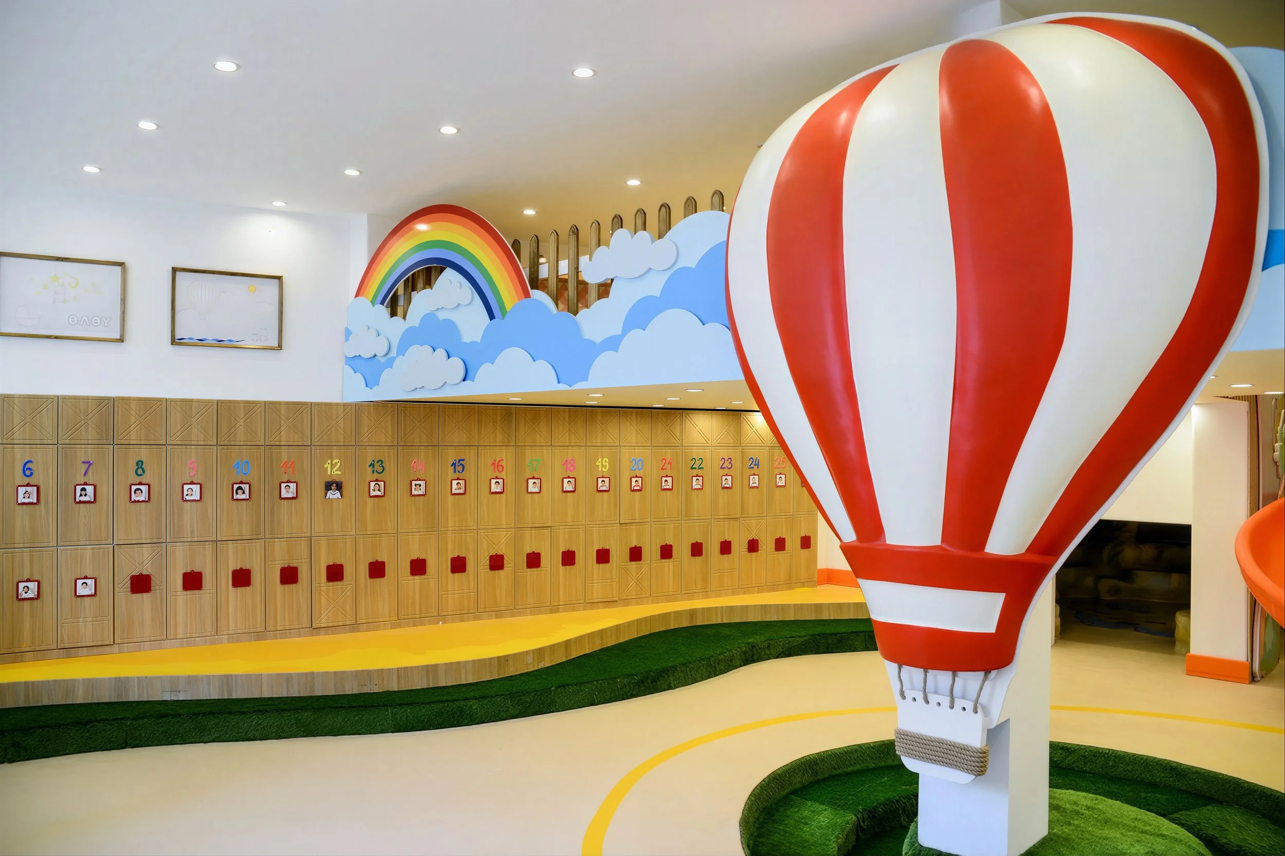 Colorful indoor space designed for children, featuring a large hot air balloon sculpture, rainbow and clouds mural, and wooden lockers with numbered photos.