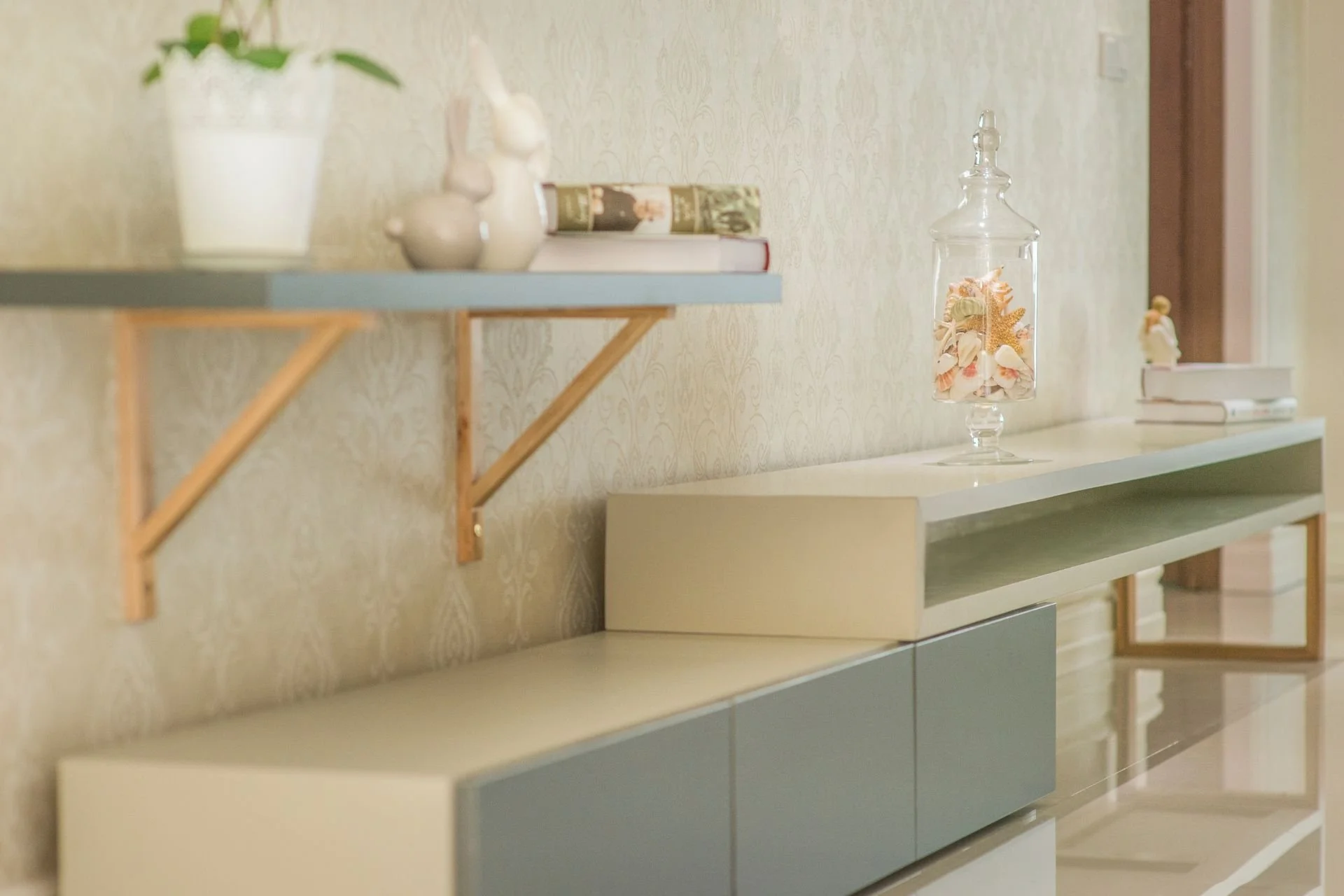 A living room with a modern white and blue TV stand, decorated with a glass jar filled with seashells and coral, and a wall-mounted shelf with ceramic rabbit figurines, books, and a potted plant.