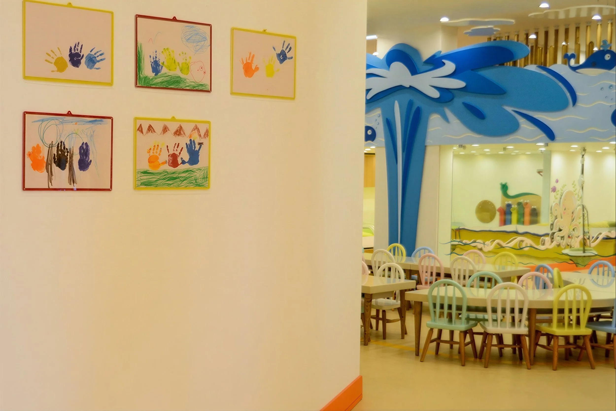 Colorful artwork of handprints and scribbles displayed on a wall in a children's play area, with a glimpse of pastel-colored chairs and play structures in the background.