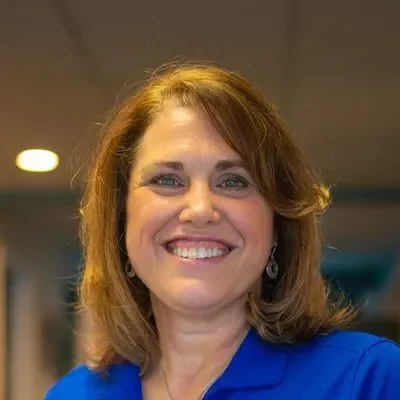 A smiling woman with shoulder-length brown hair wearing a blue top in an indoor setting.