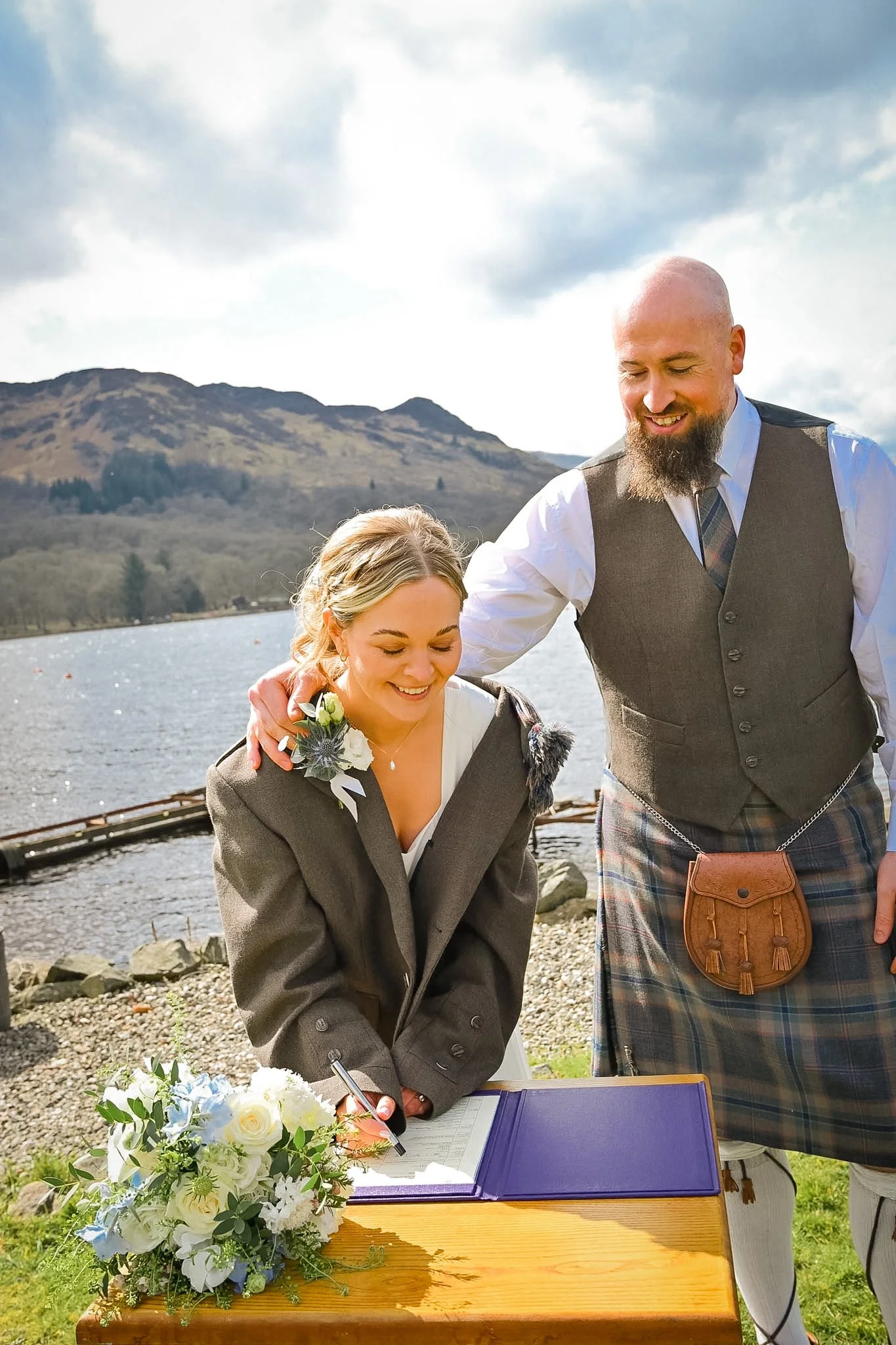 A woman in a plaid jacket signing a marriage certificate outdoors by a lake with a man in traditional Scottish attire standing beside her. They are smiling happily, and the scene is set against a backdrop of mountains and cloudy sky.