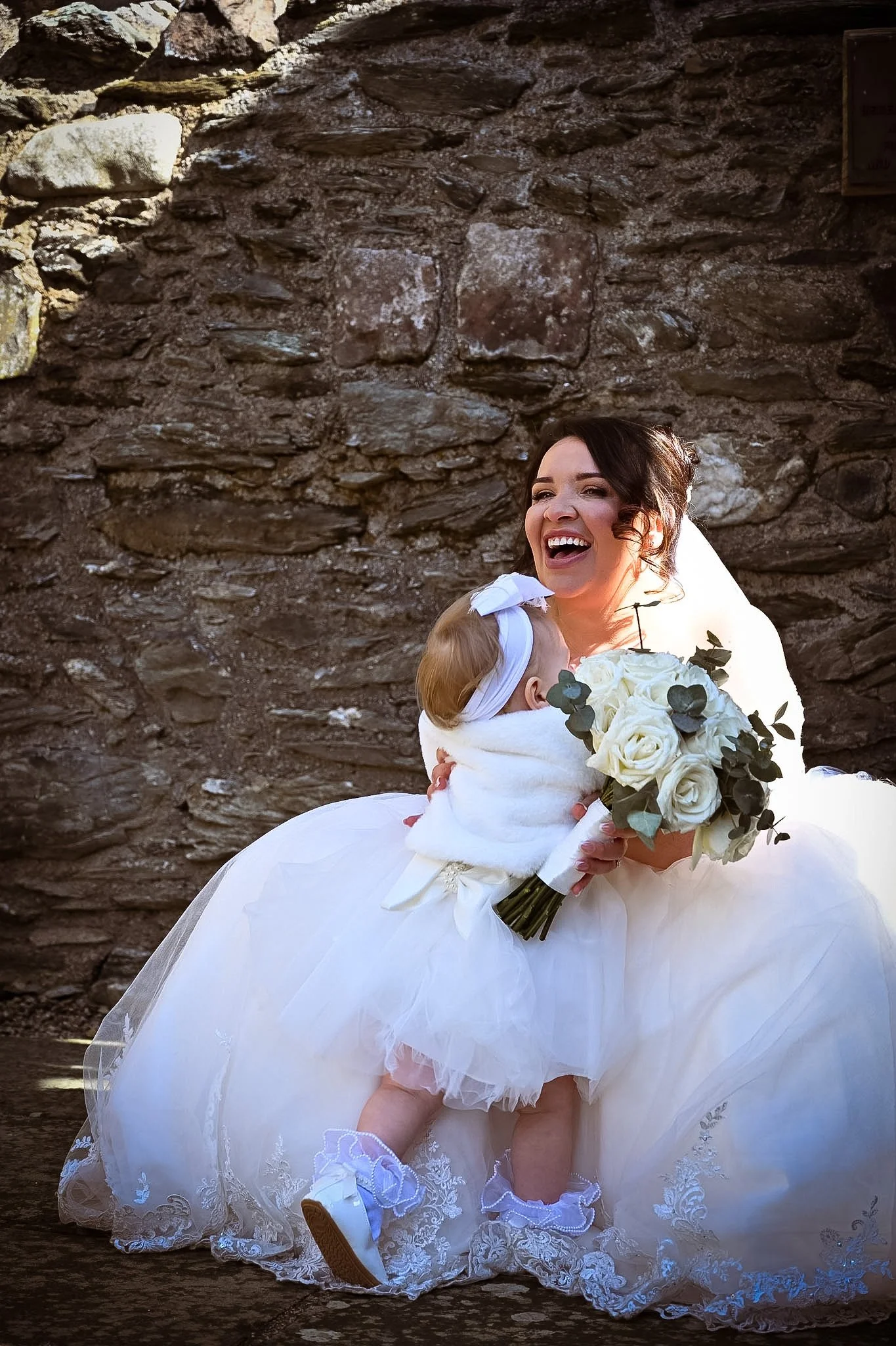 A bride in a wedding dress holding a bouquet of white roses and greenery, smiling and hugging a young girl in a white dress with lace details and a bow, outdoors with a stone wall background.